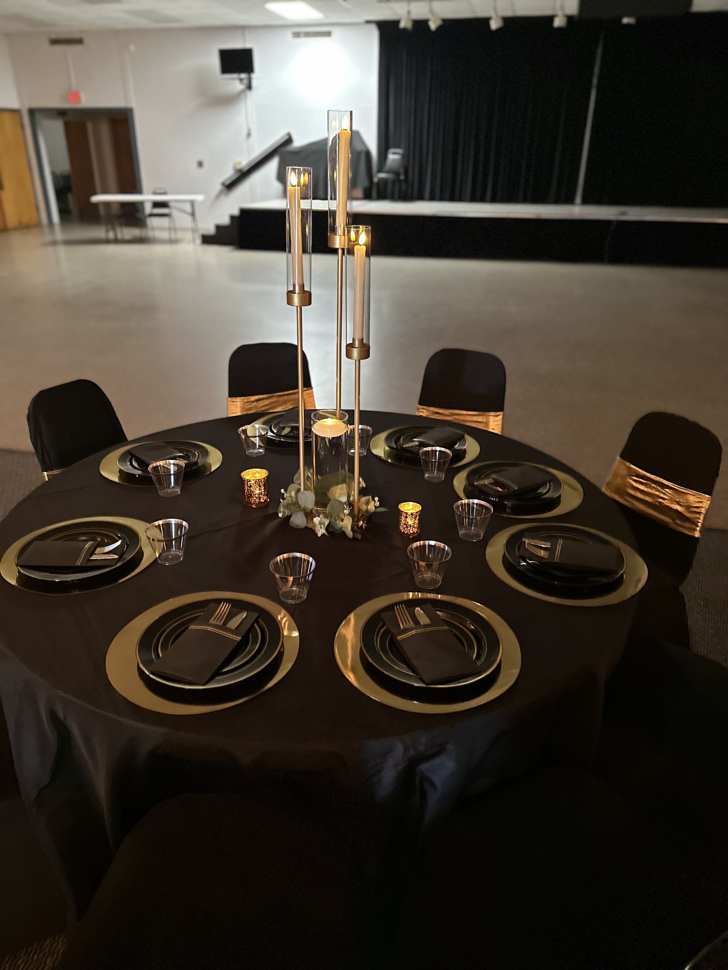 Round table decorated for a celebration or event with black tablecloth, gold and black plates, black napkins, clear cups, and a tall gold candle holder with lit candles and floral accents. The background shows a stage with black curtains and a music stand.