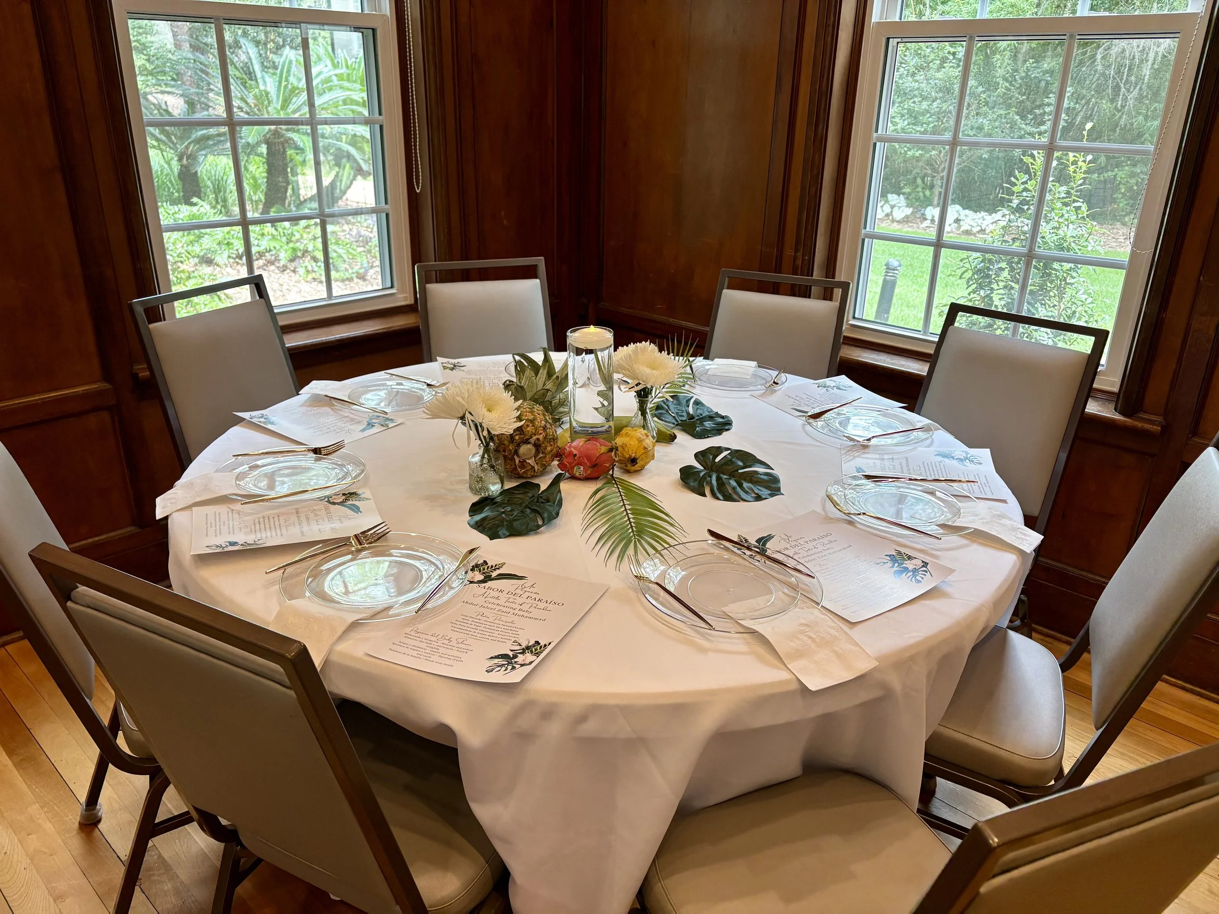 A round dining table set for a meal with white placemats, clear glass plates, and silverware. The table has a floral centerpiece with white flowers, greenery, and pineapples, and a lit candle in the middle. The room has wood-paneled walls and large windows showing greenery outside.