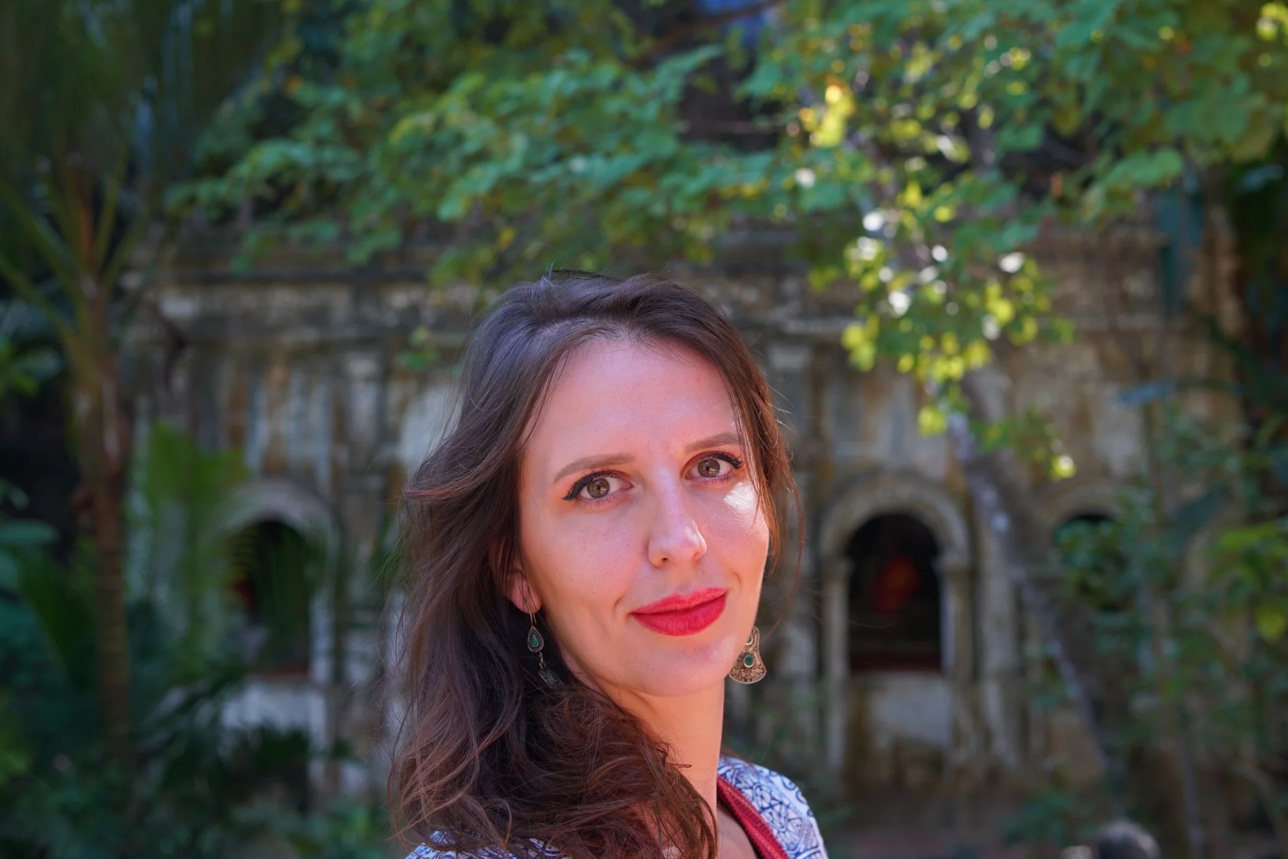 Close-up of a woman with long curly hair, wearing earrings and red lipstick, smiling outdoors with green foliage and a stone building in the background.