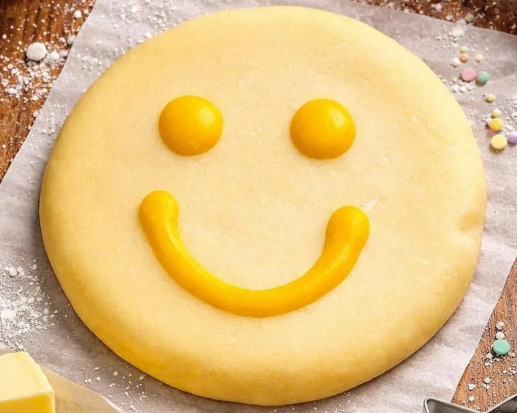 A round yellow cookie decorated with yellow frosting to resemble a smiling face, with two round dots for eyes and a curved line for a mouth.