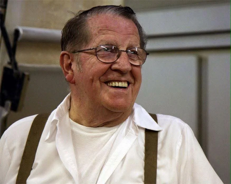 An elderly man with glasses and gray hair, smiling, wearing a white shirt and suspenders.