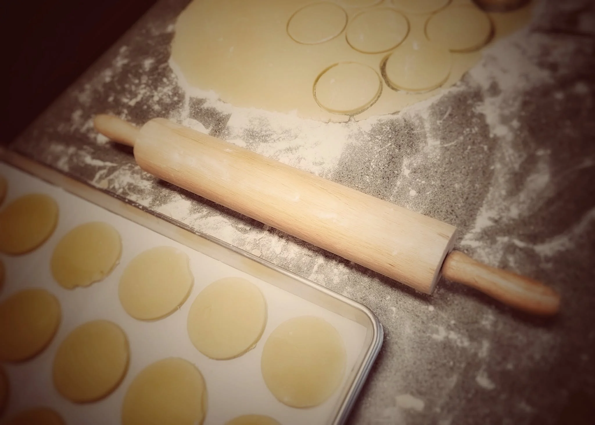 View of a kitchen countertop with a rolling pin, flour, and circular cutouts of dough on a baking sheet for making cookies or biscuits.