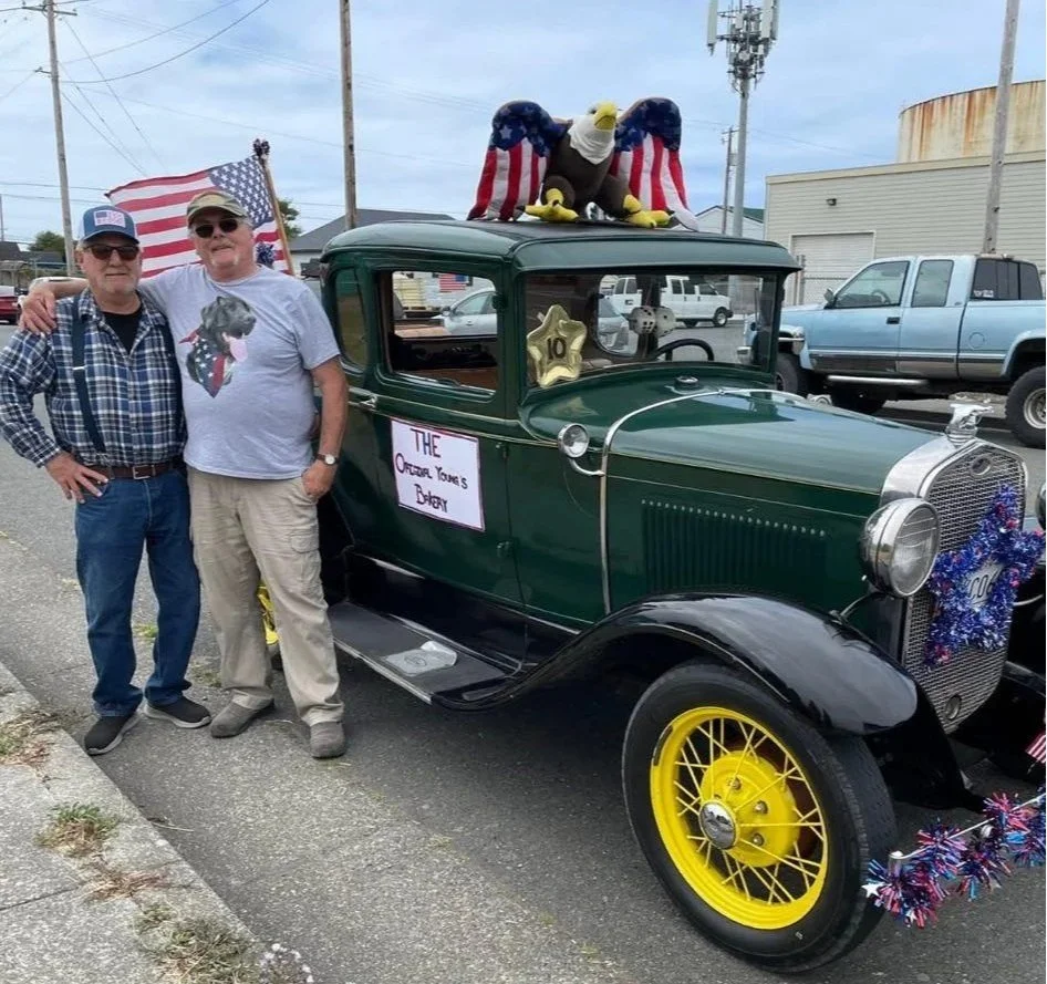 Two men standing next to a vintage green car with yellow wheels, decorated with patriotic decorations like American flags and a wreath. An eagle plush toy is on top of the car. A sign on the car reads 'The Offcial Yome's Bakery'. The background shows other vehicles and industrial buildings.