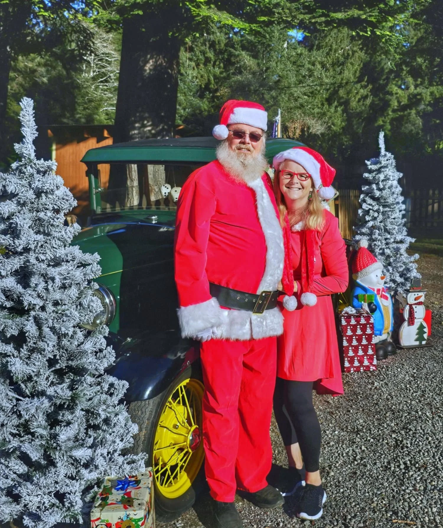 A man and woman dressed in Christmas outfits, standing outdoors near a vintage green car with yellow wheels, surrounded by Christmas decorations including snow-covered trees and gifts, with large trees and a wooden fence in the background.