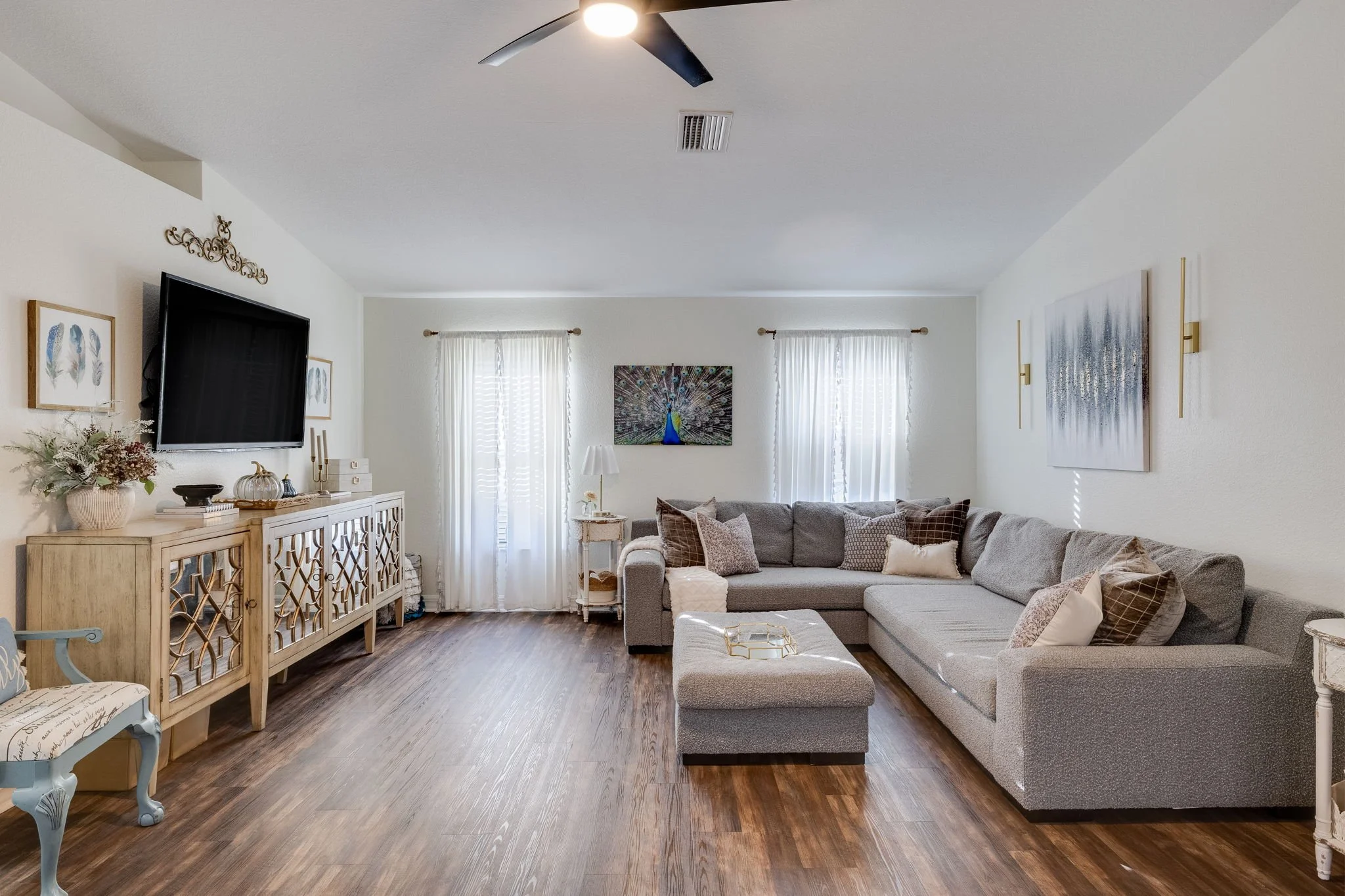 A living room with a beige sectional sofa filled with pillows, a wooden sideboard with decor, a wall-mounted TV, and framed artwork, with white curtains covering windows and wood flooring.