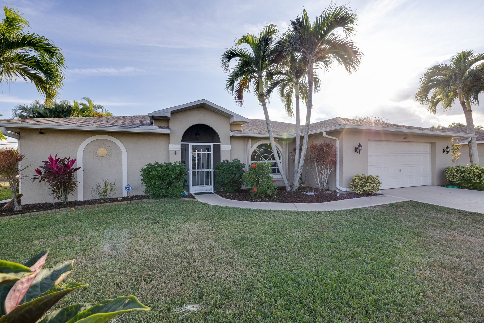 Front view of a house with a beige exterior, a two-car garage, and palm trees in the yard during daytime.