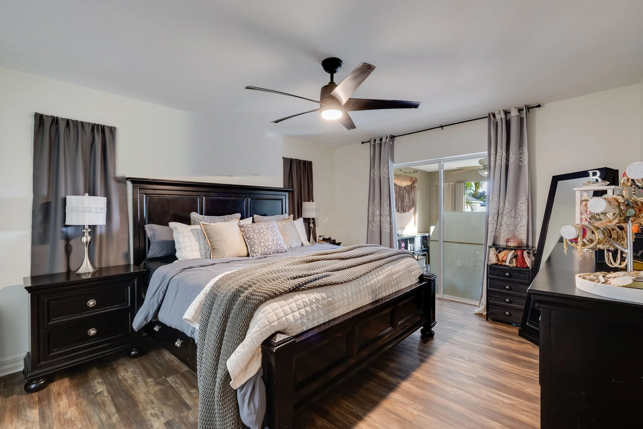 A bedroom with a dark wooden bed frame, beige bedding, throwing a gray knitted blanket over the foot, and pillows in various shades of beige and gray. There are dark nightstands with white lamps on each side, light-colored walls, and curtains covering sliding glass doors leading to a balcony with visible outdoor scenery.