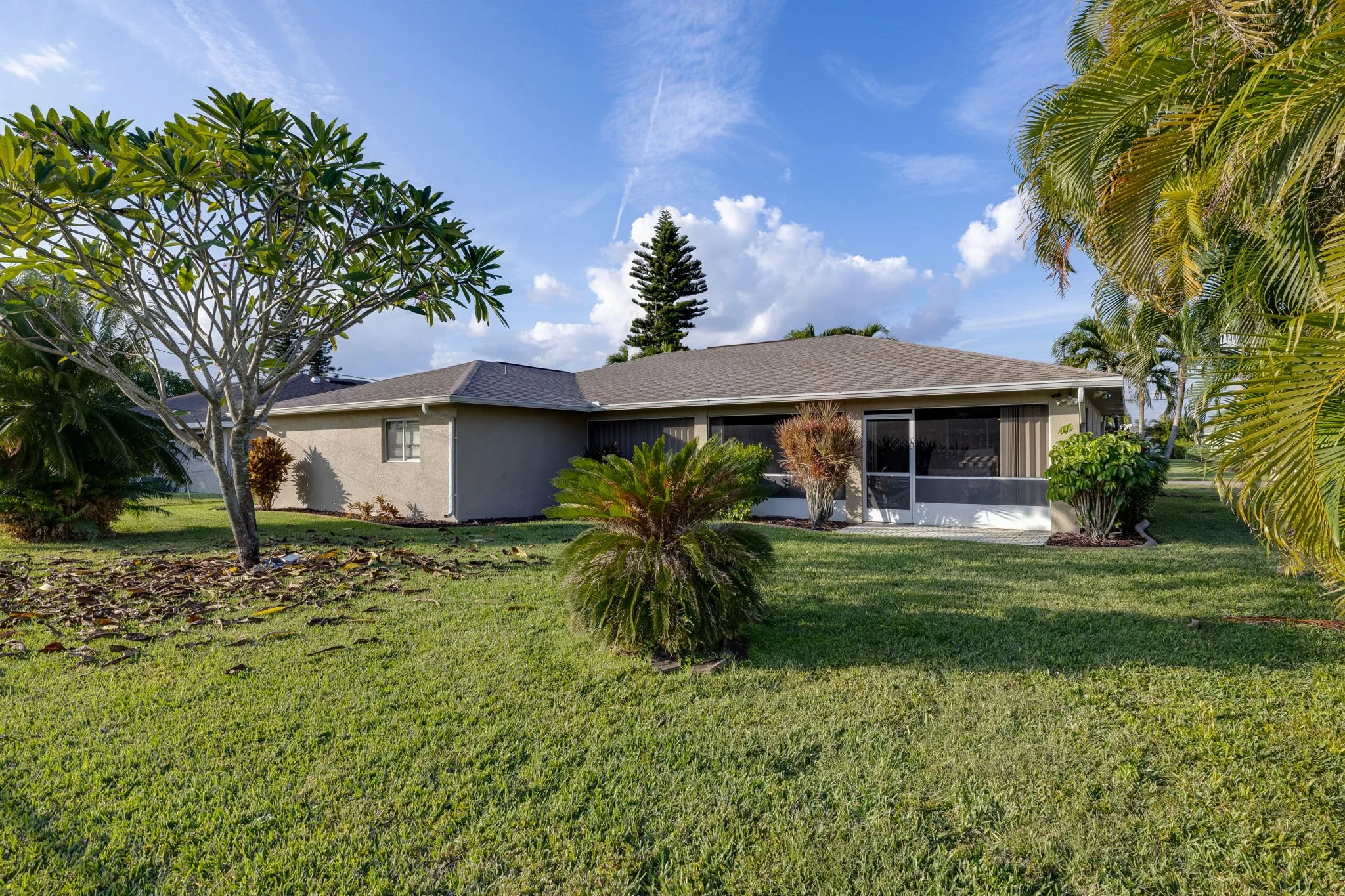 A backyard scene showing a house with a gray roof, surrounded by lush green trees and plants, under a partly cloudy sky.