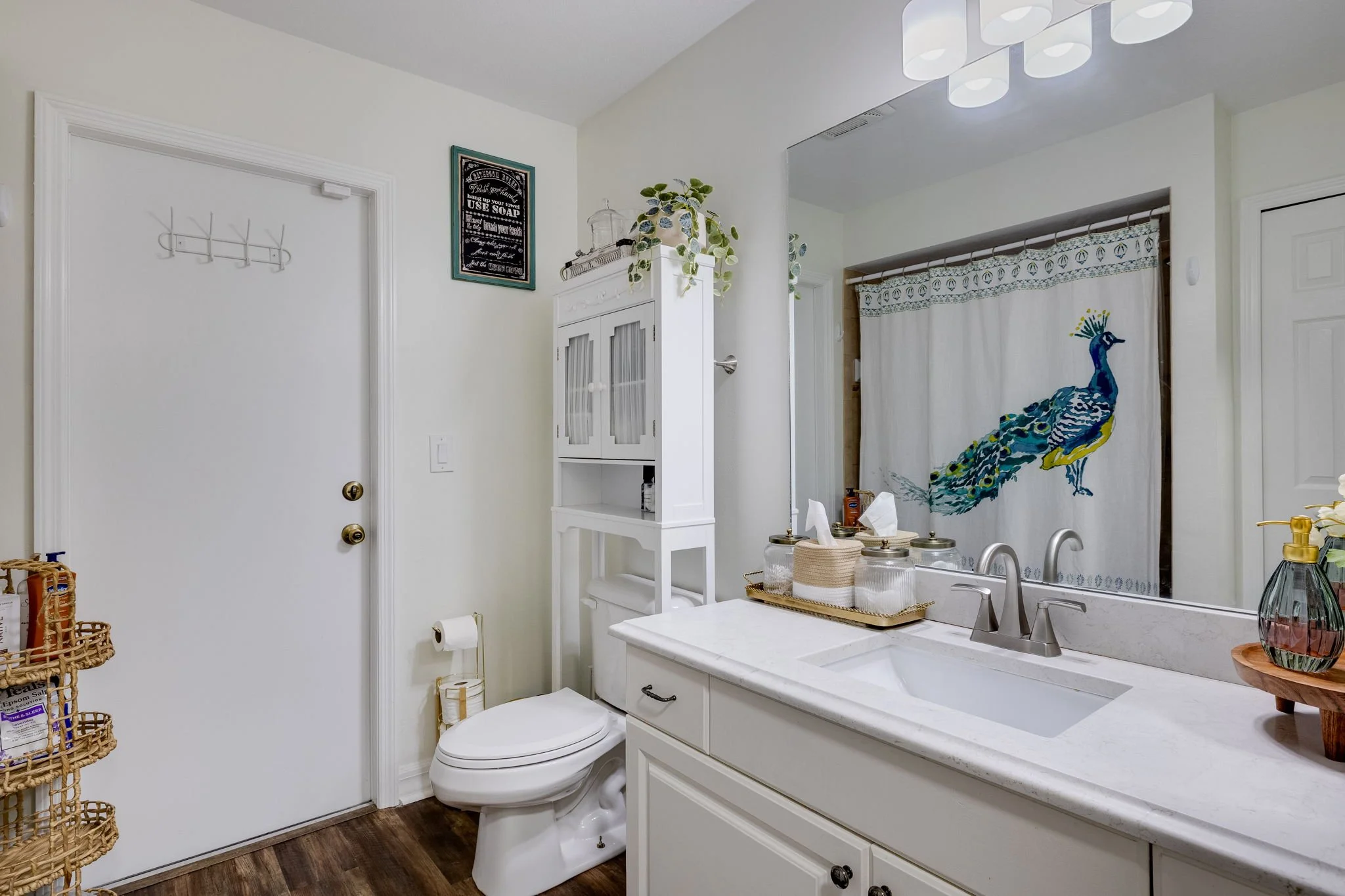 A bathroom with white walls and cabinetry, a white toilet, a white countertop with a sink, a peacock-themed shower curtain, and decorative items including a plant, a soap dispenser, and jars.