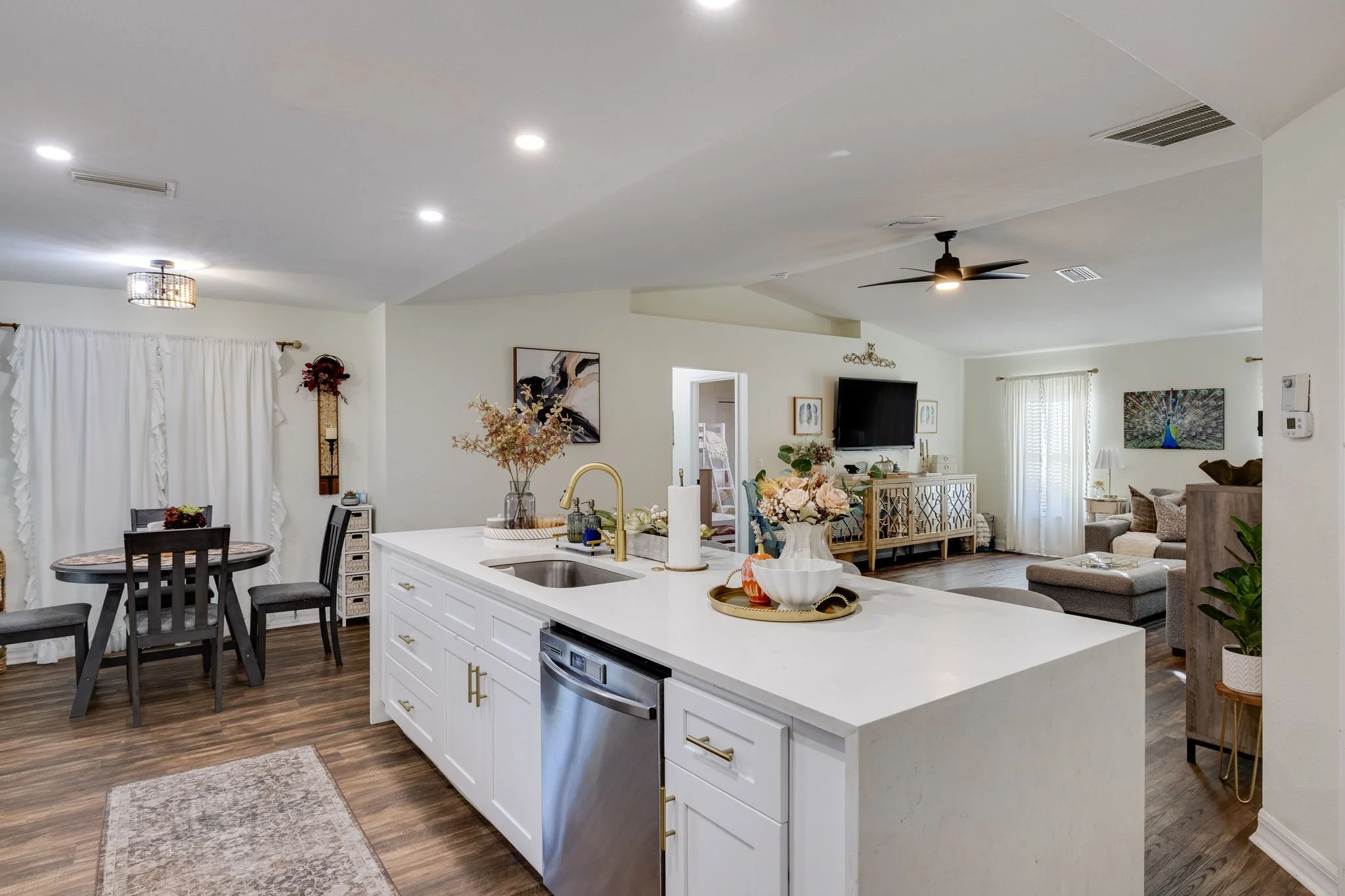 Open-concept kitchen and living room with white cabinetry, dining table, floral arrangements, wall art, ceiling fan, and natural light from windows.