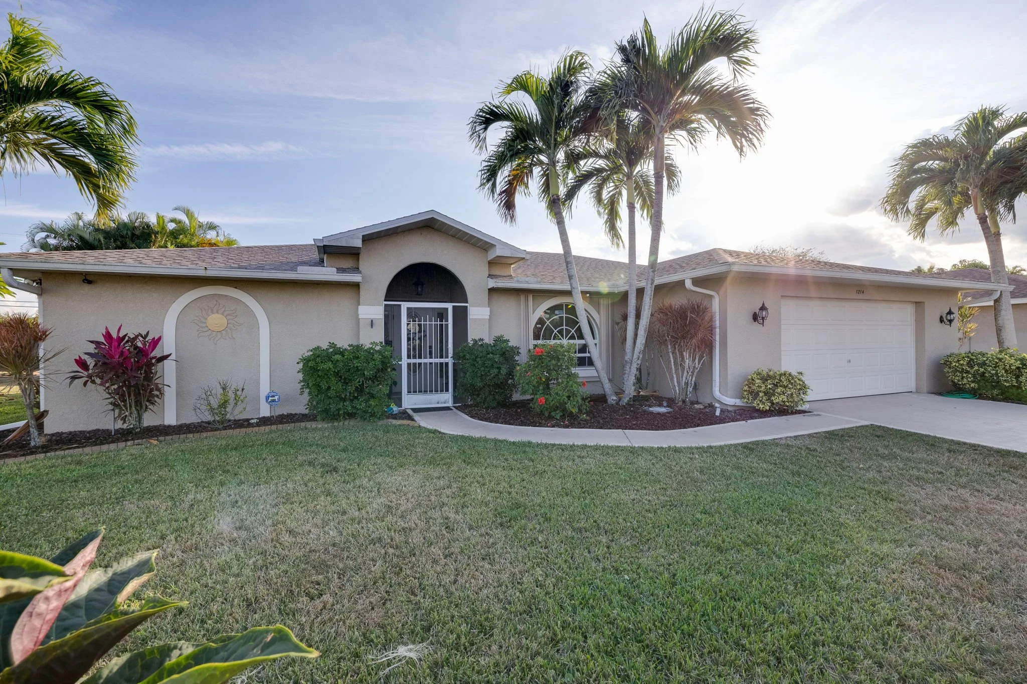 Front view of a single-story house with a well-maintained lawn, tropical palm trees, bushes, and a driveway. The house has a beige exterior with a white door and garage, and decorative sun emblem on the wall.