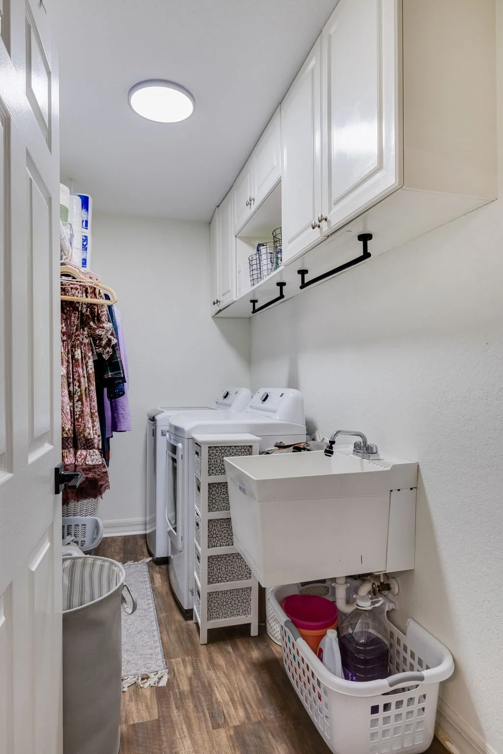 A laundry room with washing machine, dryer, utility sink, white cabinets, hanging clothes, laundry supplies, and a trash basket.