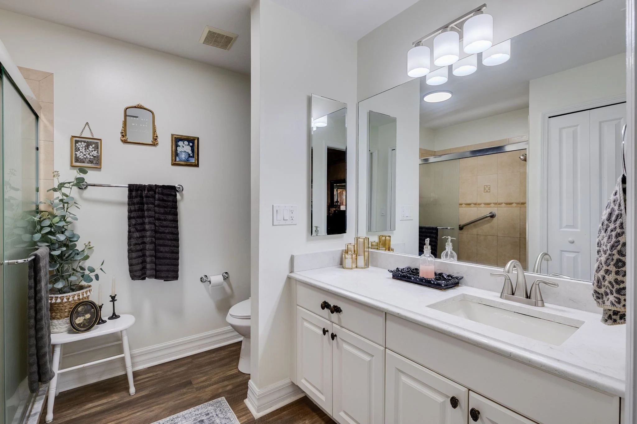 A bathroom with white cabinetry, a large mirror, and a beige tile shower stall. Decor includes framed artwork, decorative items, and plants, with wood-style flooring and modern light fixtures.