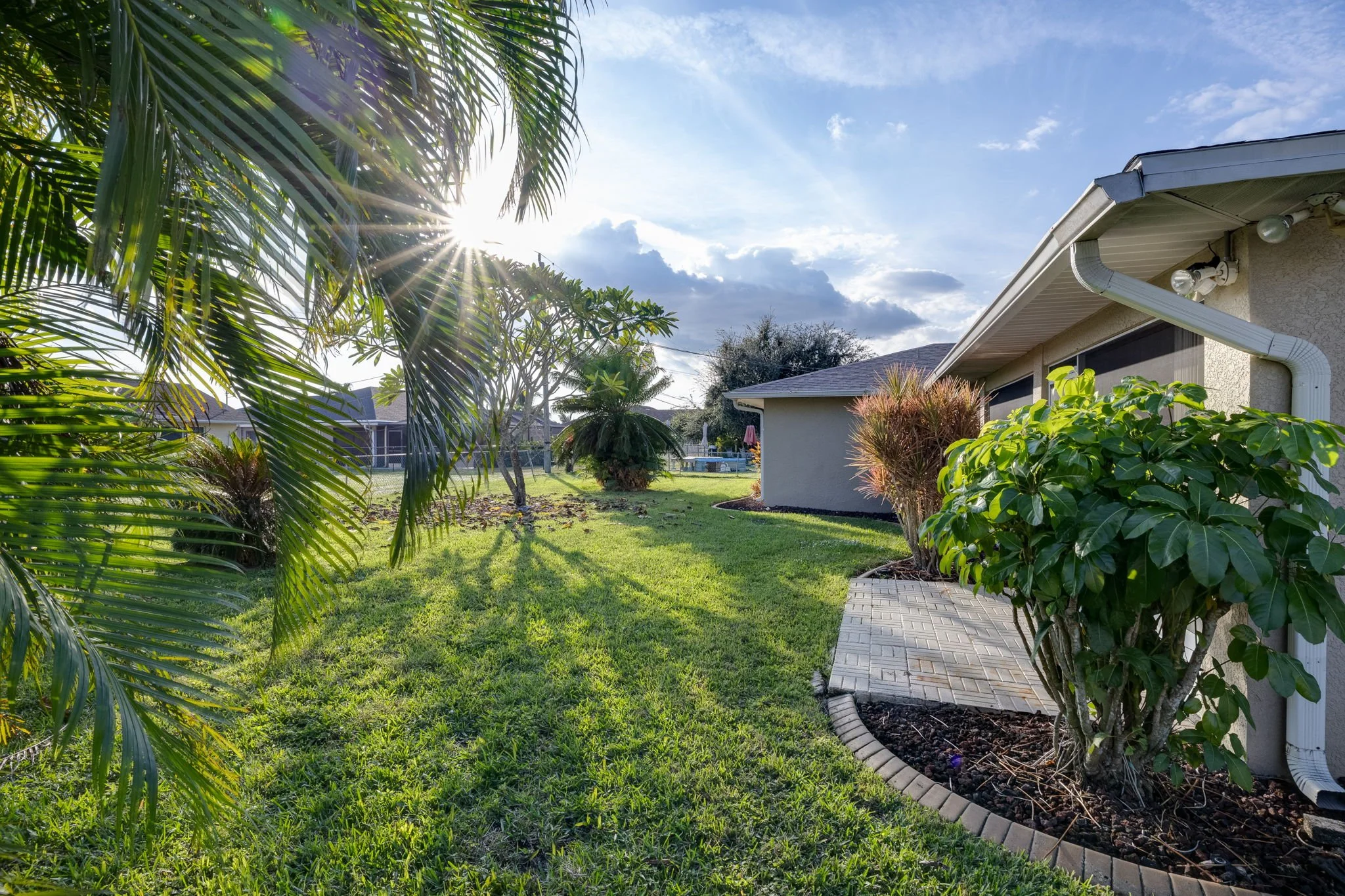 Sun shining over a backyard with green grass, palm trees, shrubs, and a house with an attached patio and gutter system.