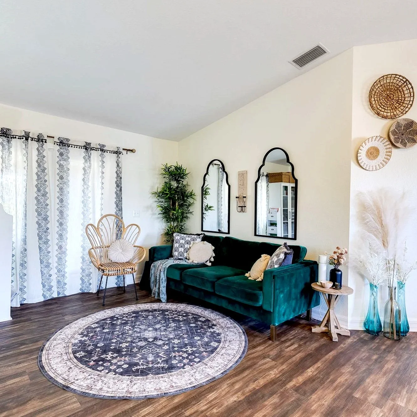 Living room with a dark green velvet sofa, decorative pillows, a round patterned rug, a wooden chair with a cushion, wall mirrors, a large potted plant, and decorative baskets on the wall. Floor-length curtains with a geometric pattern hang on a window.