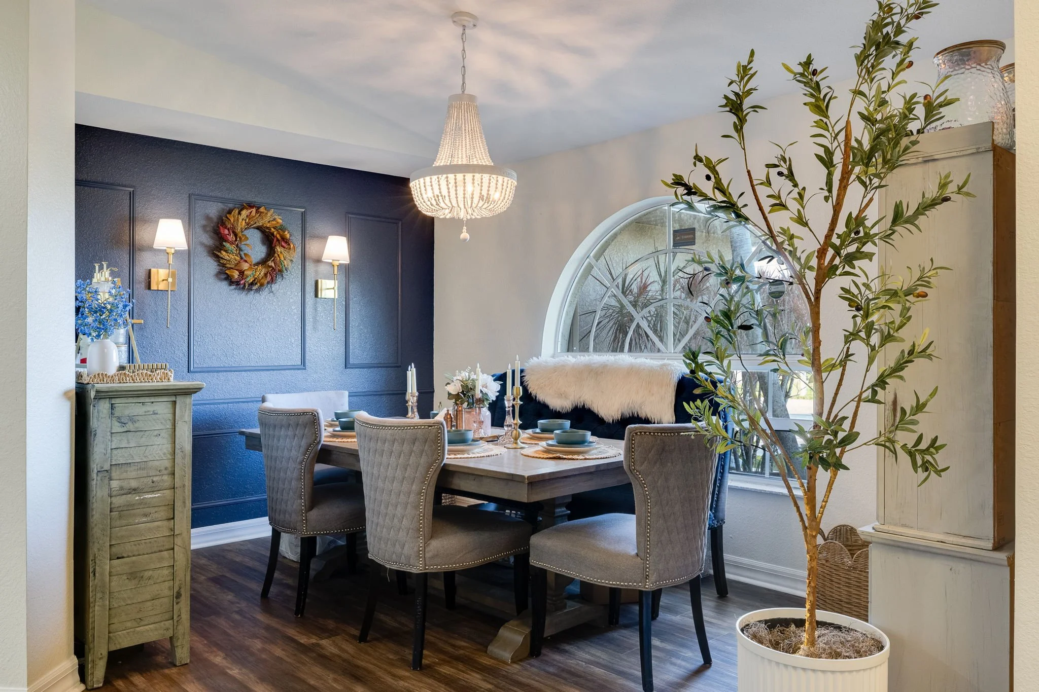 A dining room with a natural wood table set with a centerpiece of flowers and candles, beige chairs with nailhead trim, a boat-shaped window, a large leafy plant in a white planter, a black wall with a decorative wreath, and a white chandelier hanging from the ceiling.