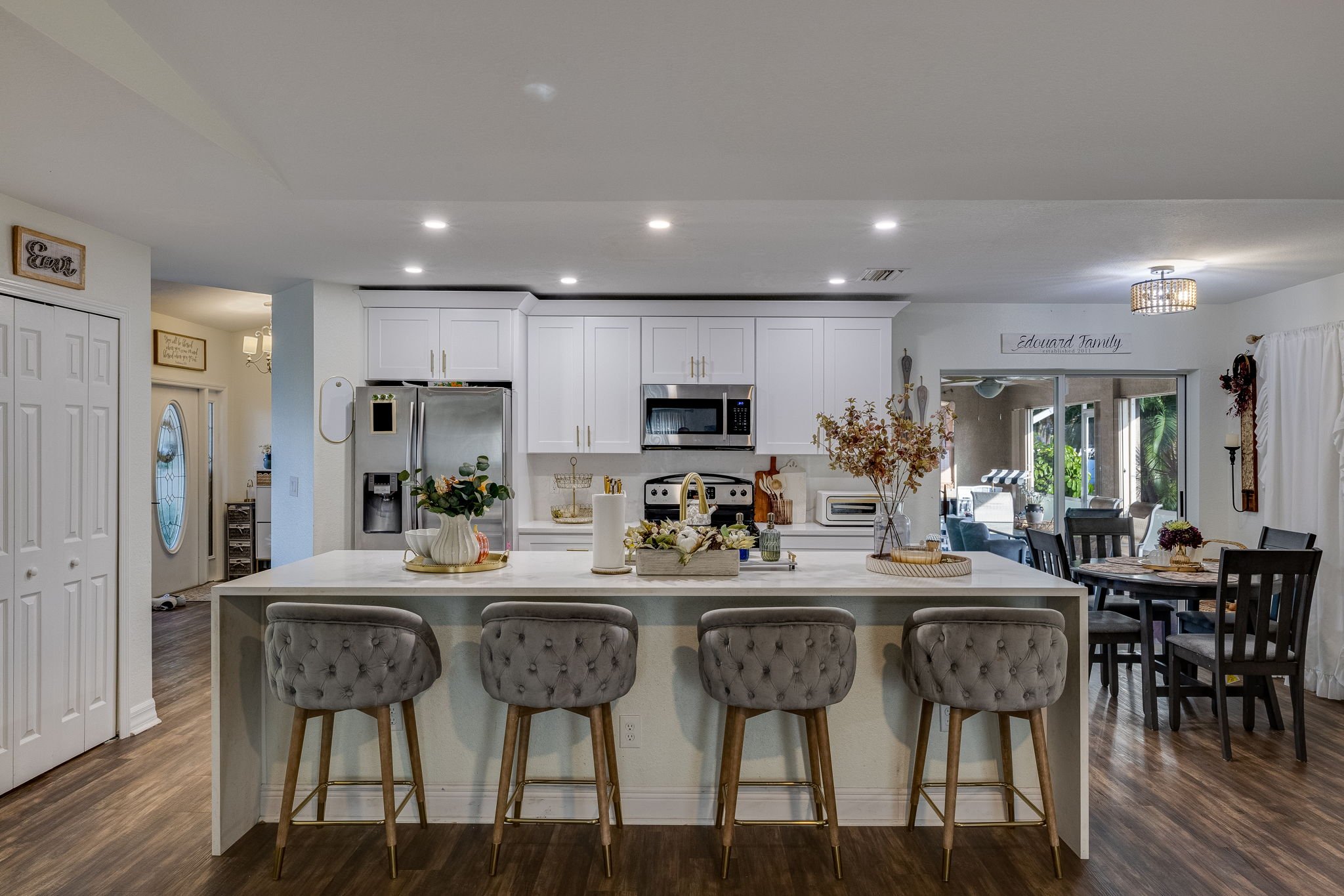 A modern open-plan kitchen with white cabinets, stainless steel appliances, and a large island. The island has four plush beige upholstered bar stools with tufted backs. Decorative plants, jars, and a paper towel roll are placed on the island. In the background, there is a dining area with a table and chairs near a sliding glass door with curtains, and a view of outdoor greenery.