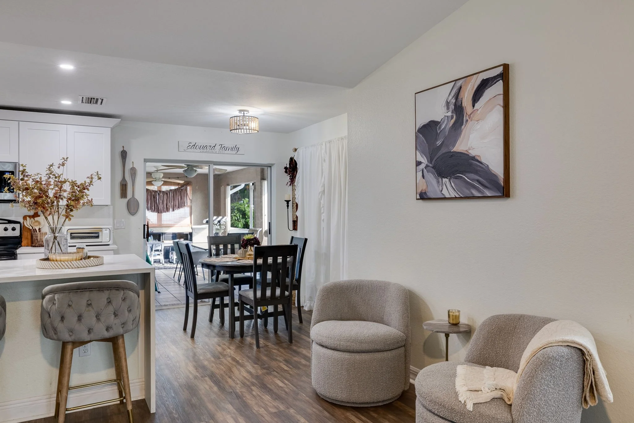 A cozy living area and dining space in a home with white walls, a modern painting, a dining table with chairs, and a view into an outdoor patio area.