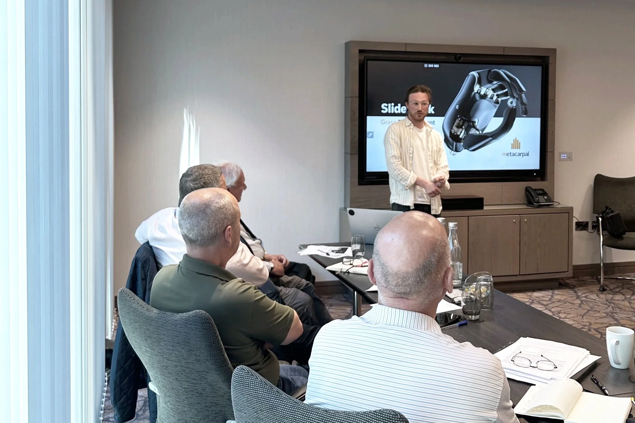 A man with glasses and a white shirt standing in front of a presentation screen with an image of a robotic hand and the text 'Slide deck'. Several people seated around a conference table are listening, with notebooks, papers, and glasses of water in front of them.