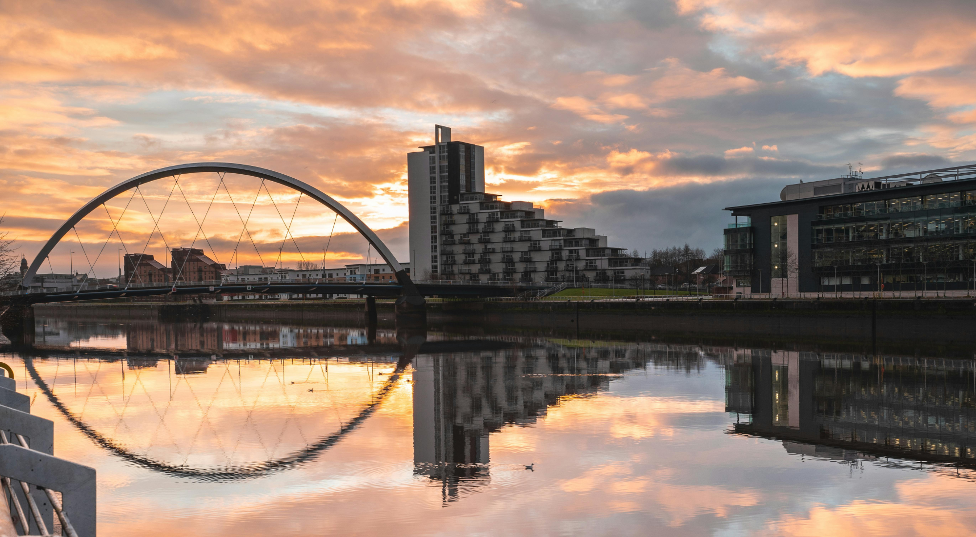 Sunset over a cityscape with modern buildings, a bridge with an arch and cables, and a calm river reflecting the sky and structures.