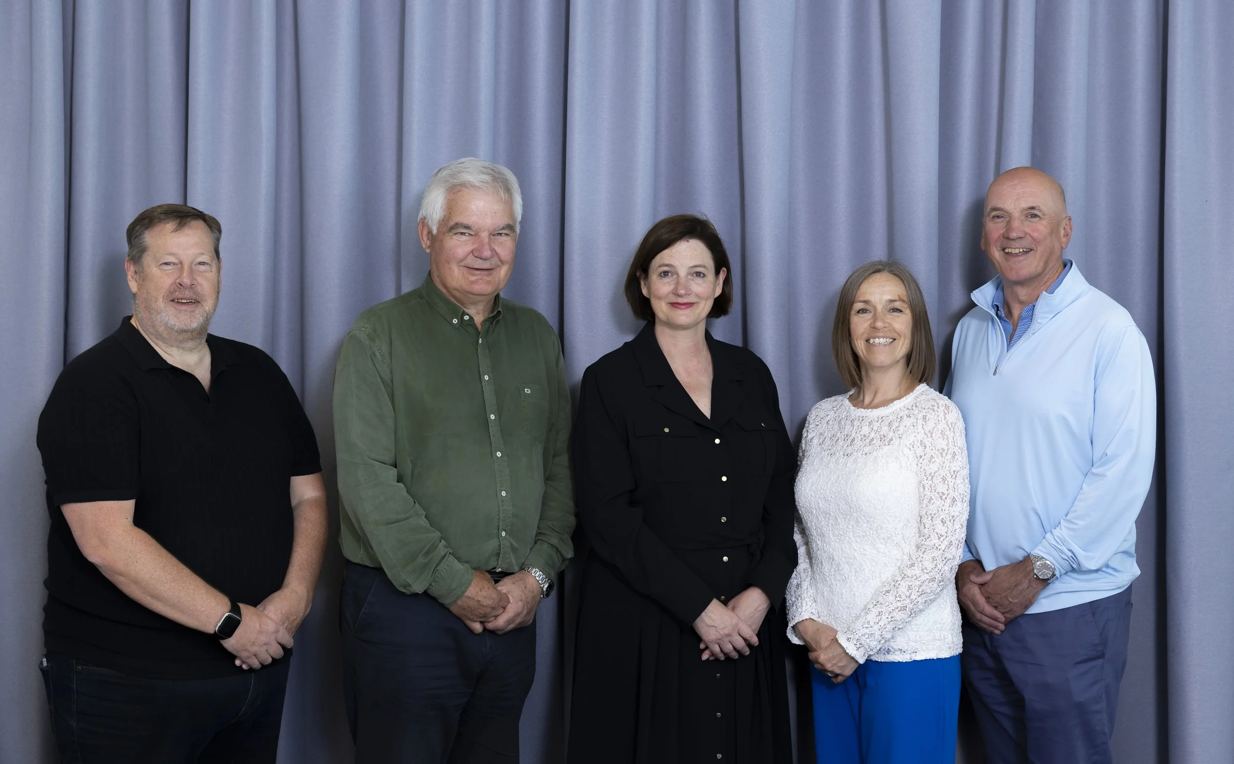 Group of six adults standing in front of a blue curtain, smiling for the camera.