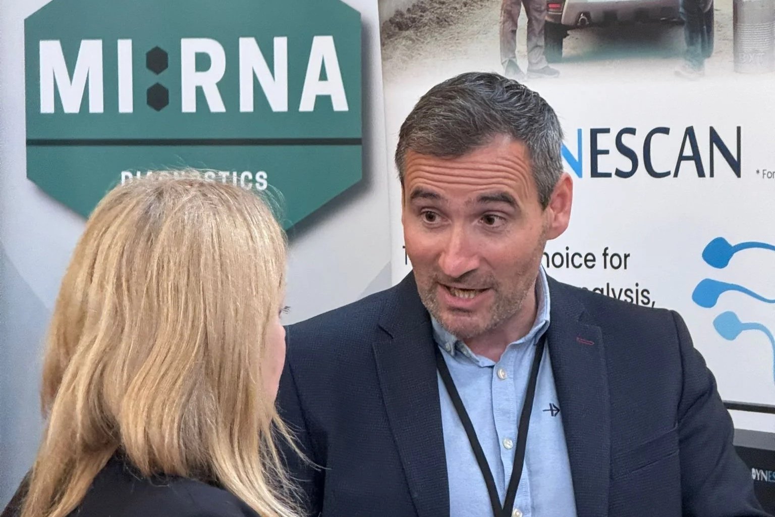 A man and a woman are having a conversation at a professional event. The man has short gray hair, a beard, and is wearing a dark blazer with a light blue shirt. There are informational banners behind them with text referencing MI:RNA Diagnostics and NESCAN.