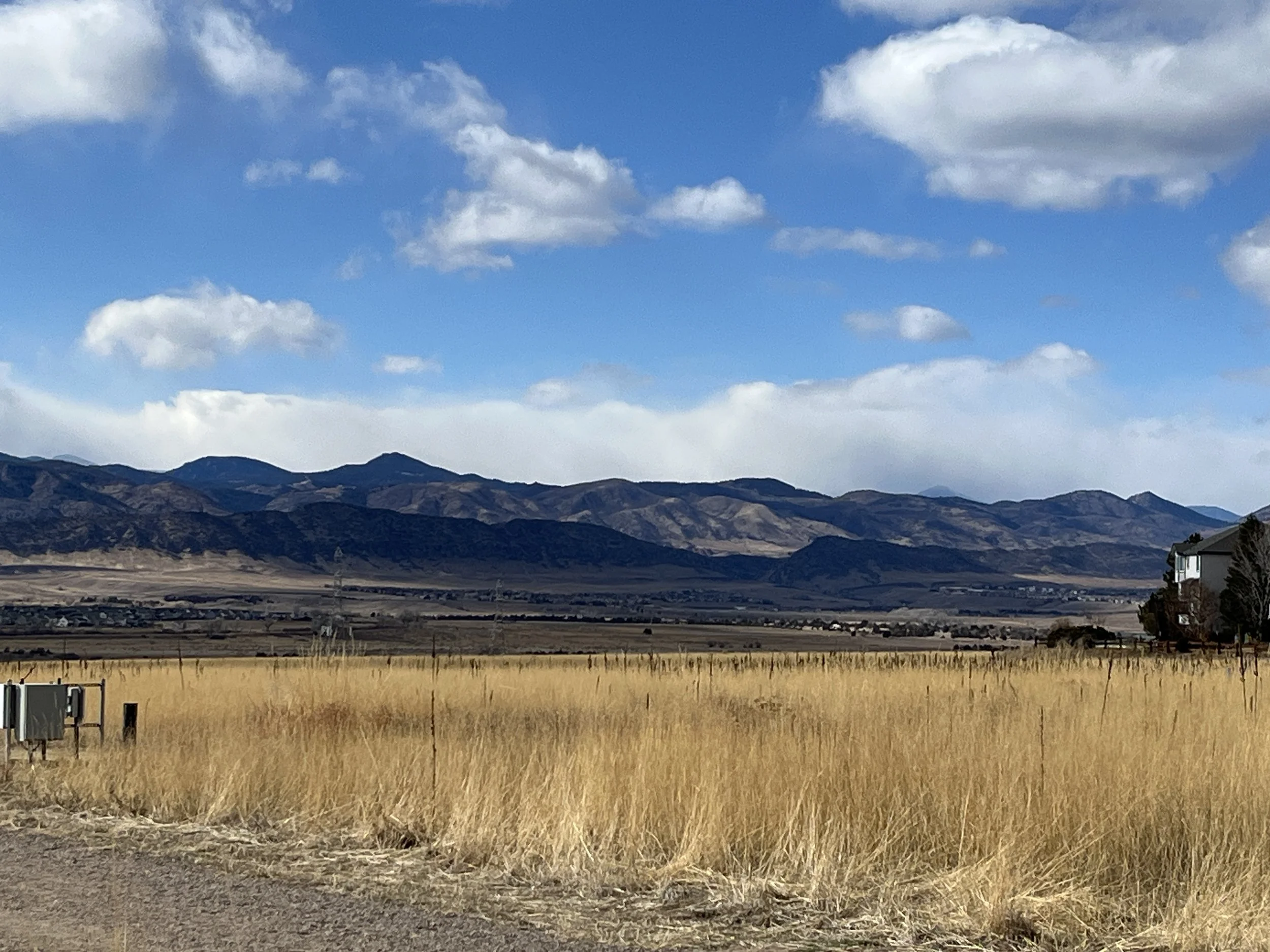View of a landscape with golden field in the foreground, utility boxes, mountains in the background, and partly cloudy sky.