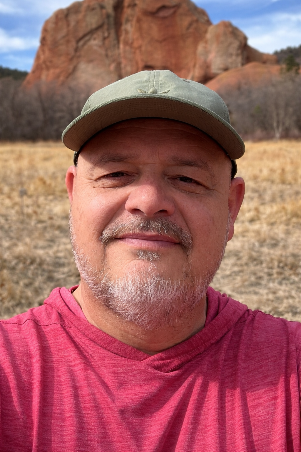 A man taking a selfie outdoors with a large red rock formation in the background, wearing a green cap and a red shirt.