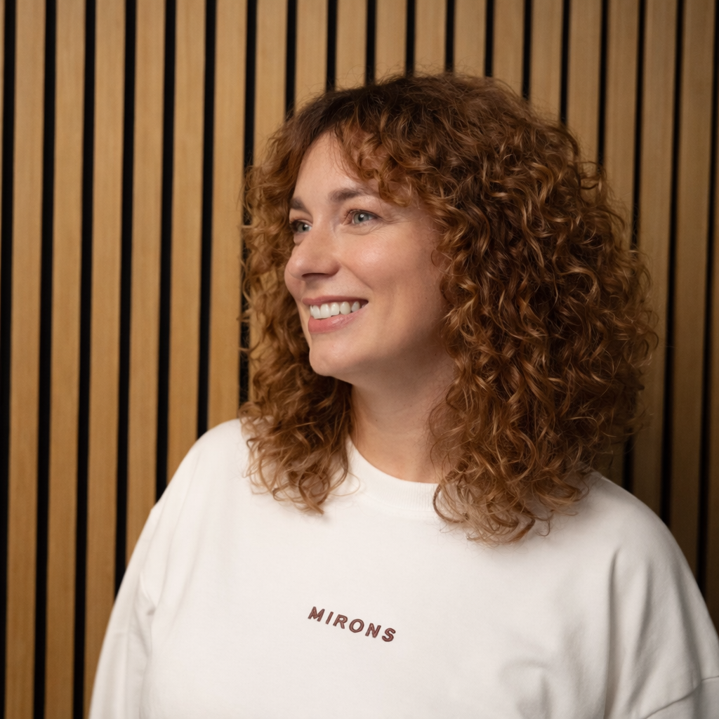 A woman with curly red hair smiling, wearing a white t-shirt with the word 'MIRONS' printed on it, standing in front of a wooden slat background.