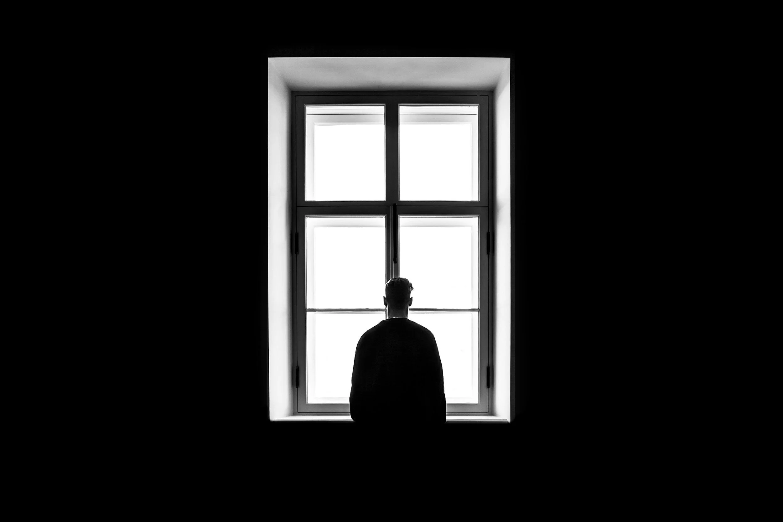 A person standing in front of a bright window, looking outside, inside a dark room.