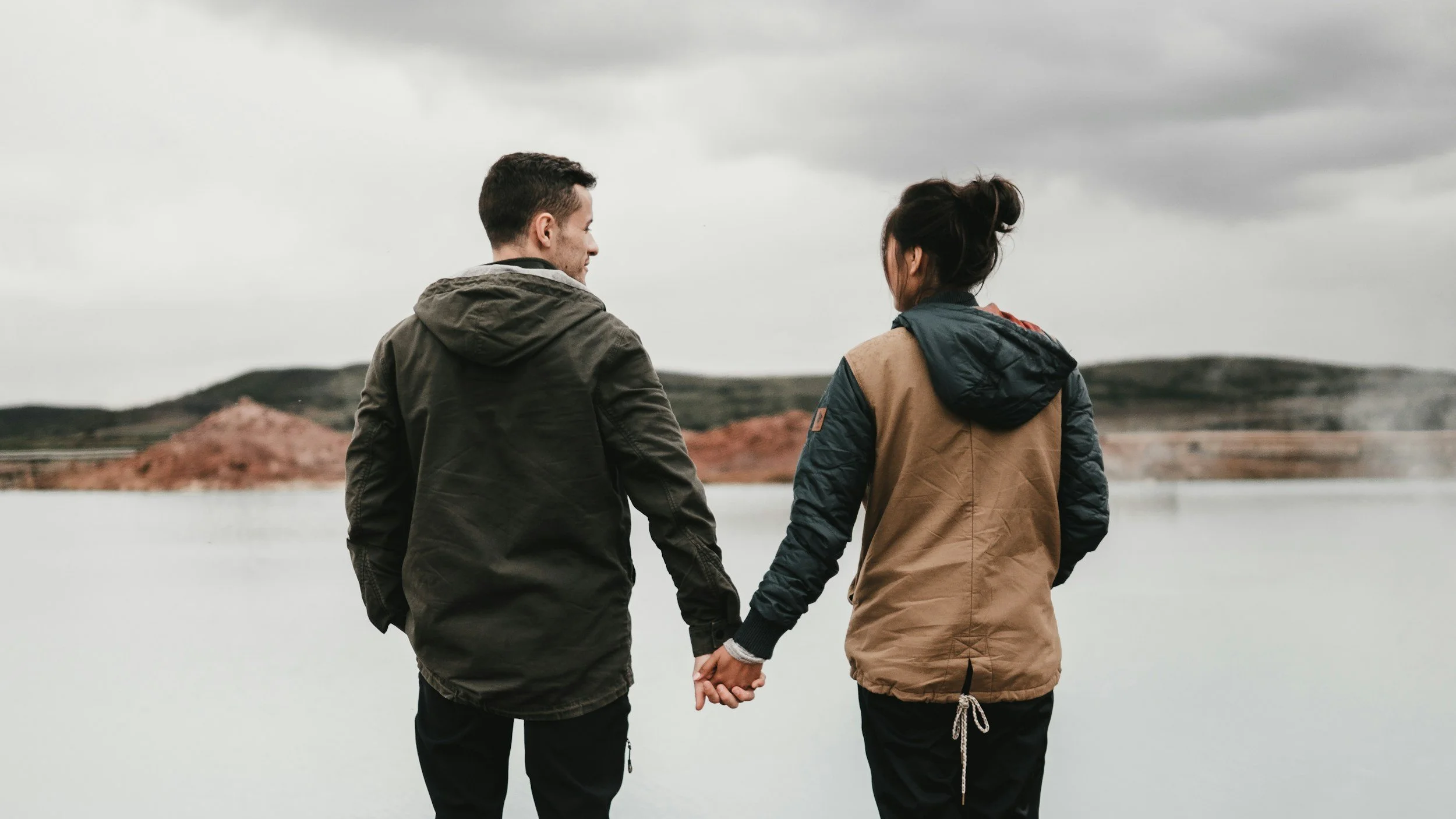 A couple holding hands and walking by a body of water under a cloudy sky.