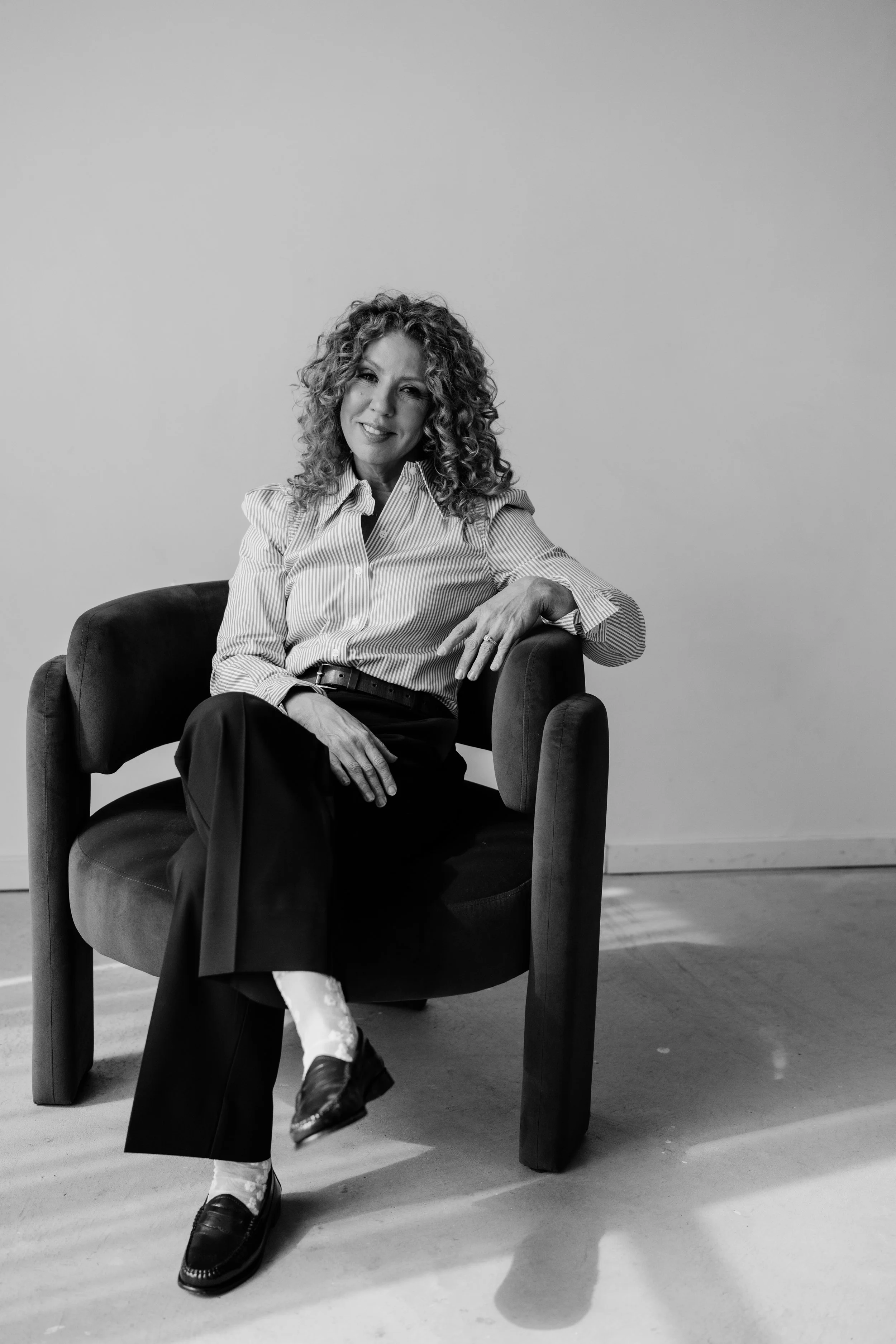 Black and white photo of a woman with curly hair sitting on a chair, smiling, wearing a striped shirt, black pants, white socks, and black shoes, in front of a plain wall.