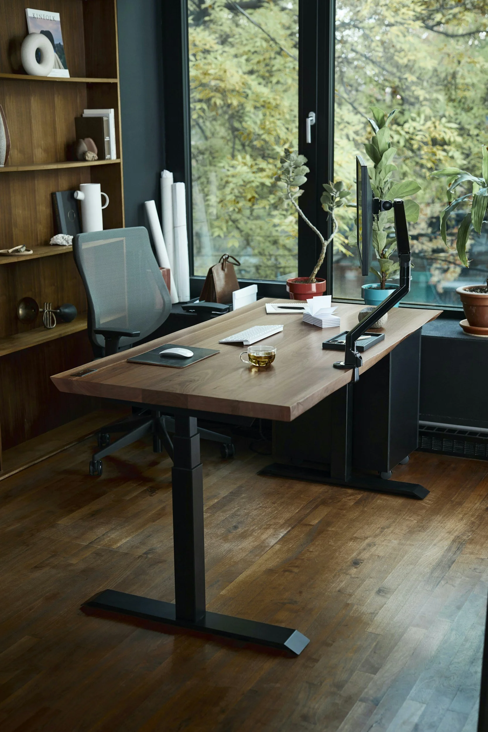 An office with a wooden desk, ergonomic chair, potted plants, computer monitor, keyboard, and mouse, near a large window with trees outside.