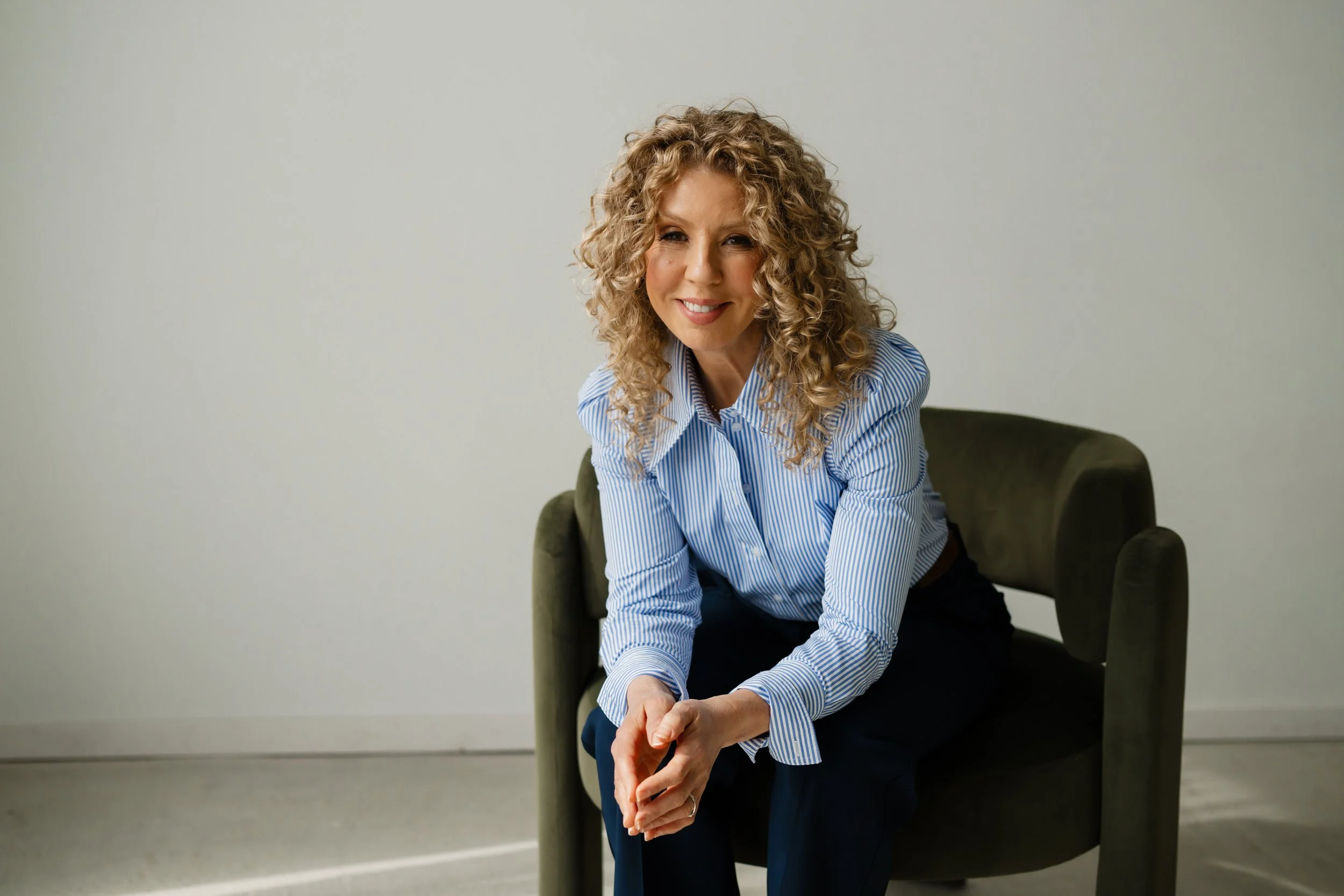 A woman with curly blonde hair smiling, wearing a striped blue and white shirt, sitting on a dark green chair against a plain light-colored wall.