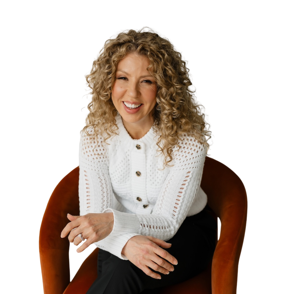 A woman with curly blonde hair smiling, sitting in a brown chair, wearing a white sweater with buttons and black pants.