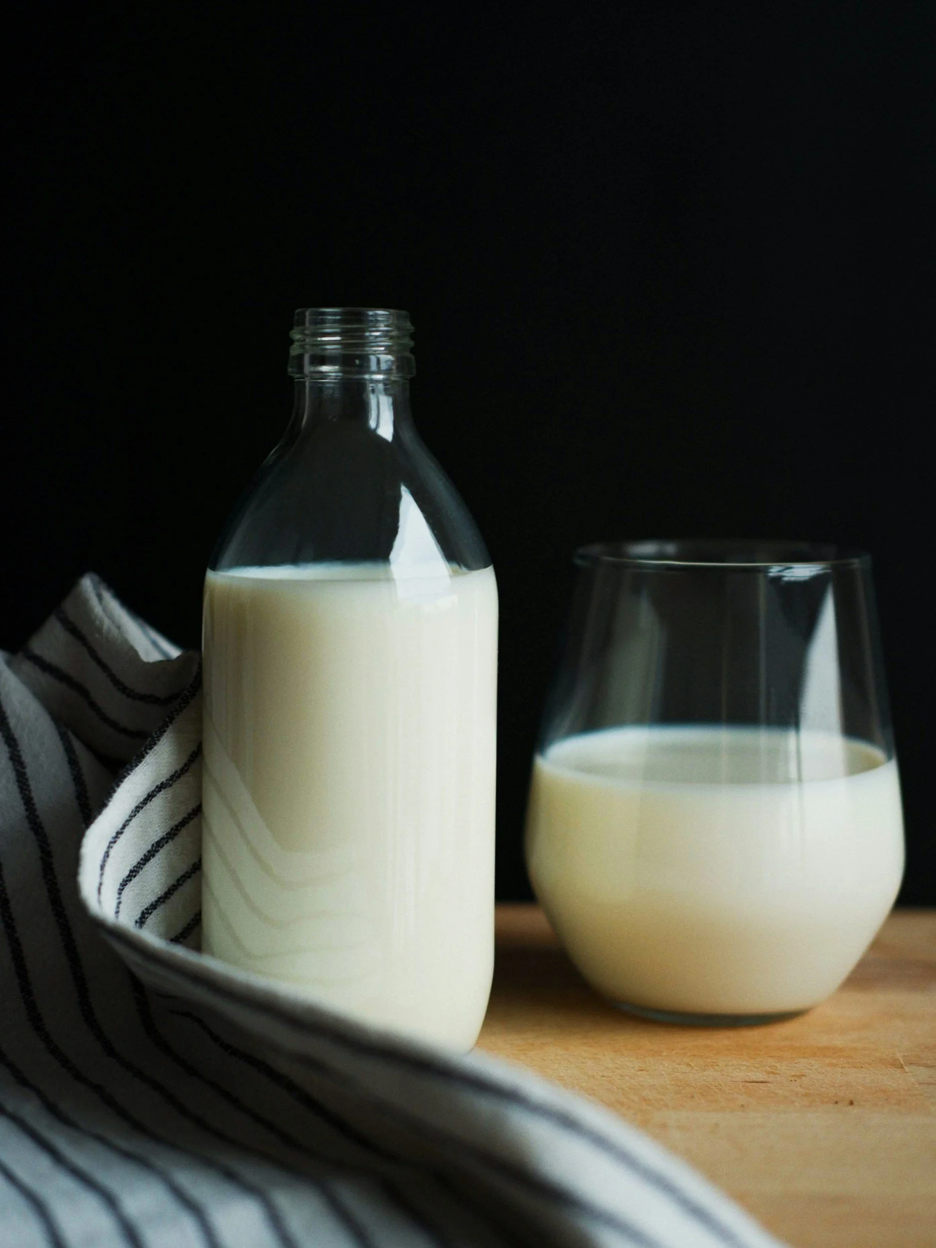 A clear glass bottle filled with milk next to a glass of milk on a wooden surface, with a black background.