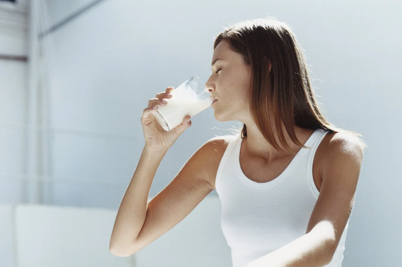 A woman with brown hair and a white tank top is drinking a glass of milk.
