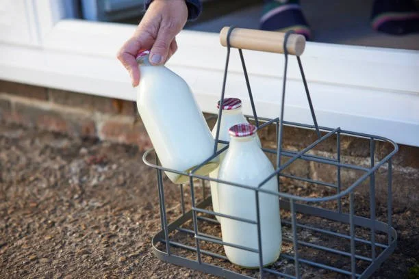 Person placing a glass bottle of milk into a metal outdoor storage basket on the ground next to a house window.