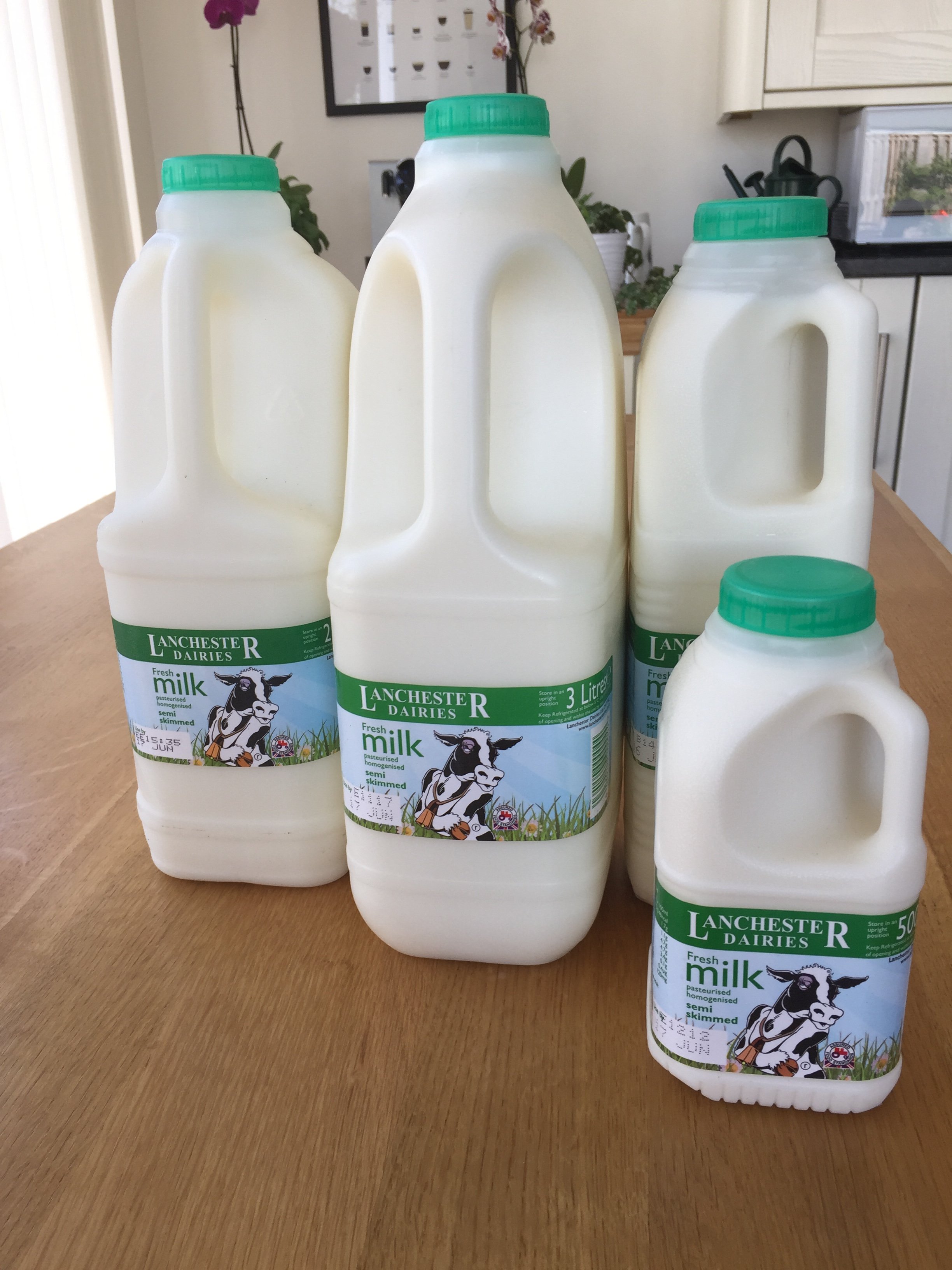 Six bottles and one container of Lancaster Dairies milk, with green caps, displayed on a wooden table in a kitchen.