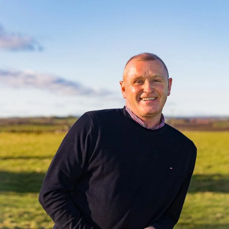 A man smiling outdoors in a grassy field with a blue sky and some clouds, wearing a black sweater and a collared shirt.