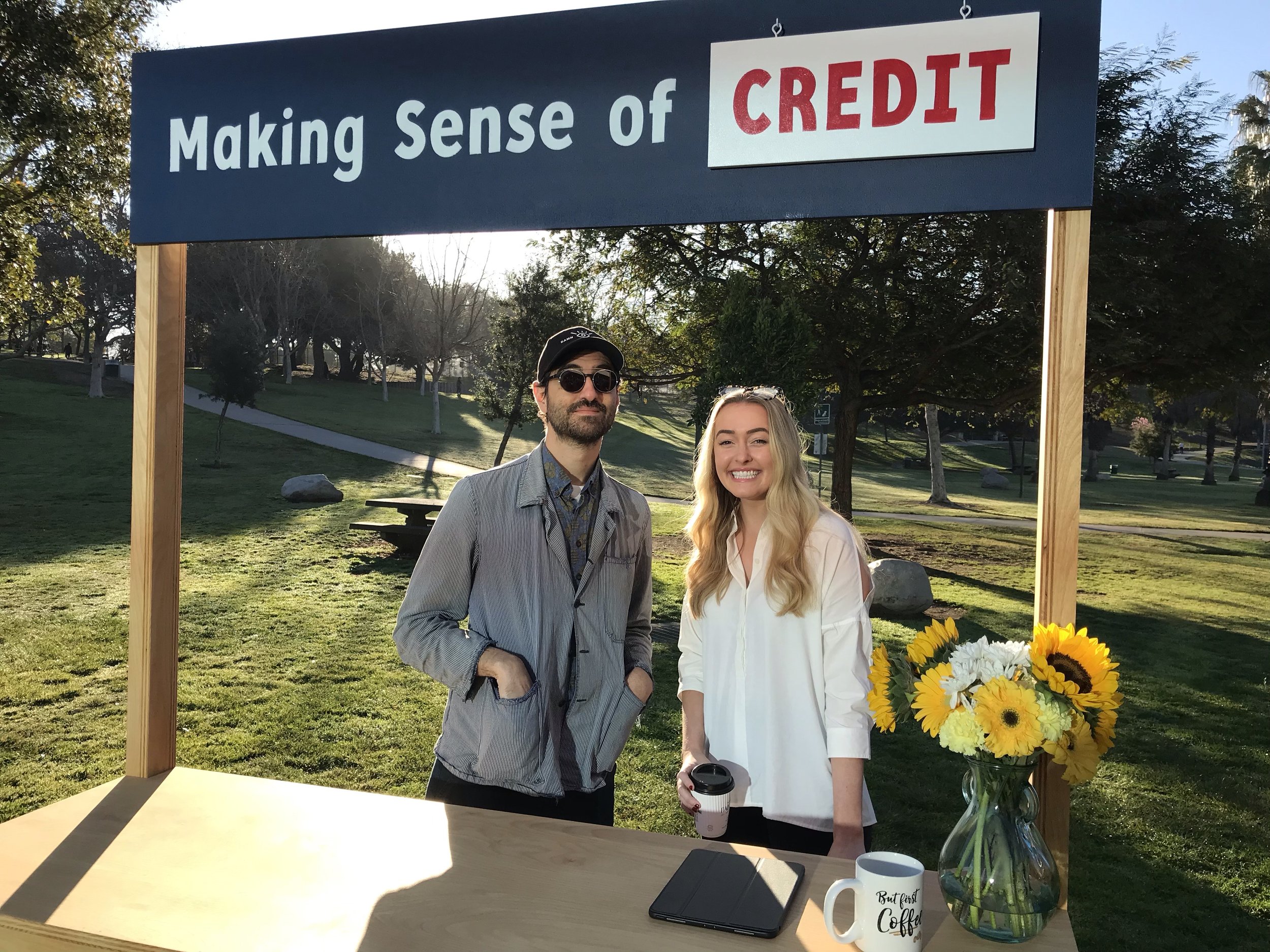 Two people standing outside under a sign that says "Making Sense of Credit". A man with a beard wearing sunglasses, a striped blazer, and a collared shirt, and a smiling woman with long blonde hair wearing a white blazer, holding a bouquet of sunflowers and white flowers. A table in front of them has a black notebook, a white mug with black text, a disposable coffee cup, and a vase with a bouquet of sunflowers and white flowers.