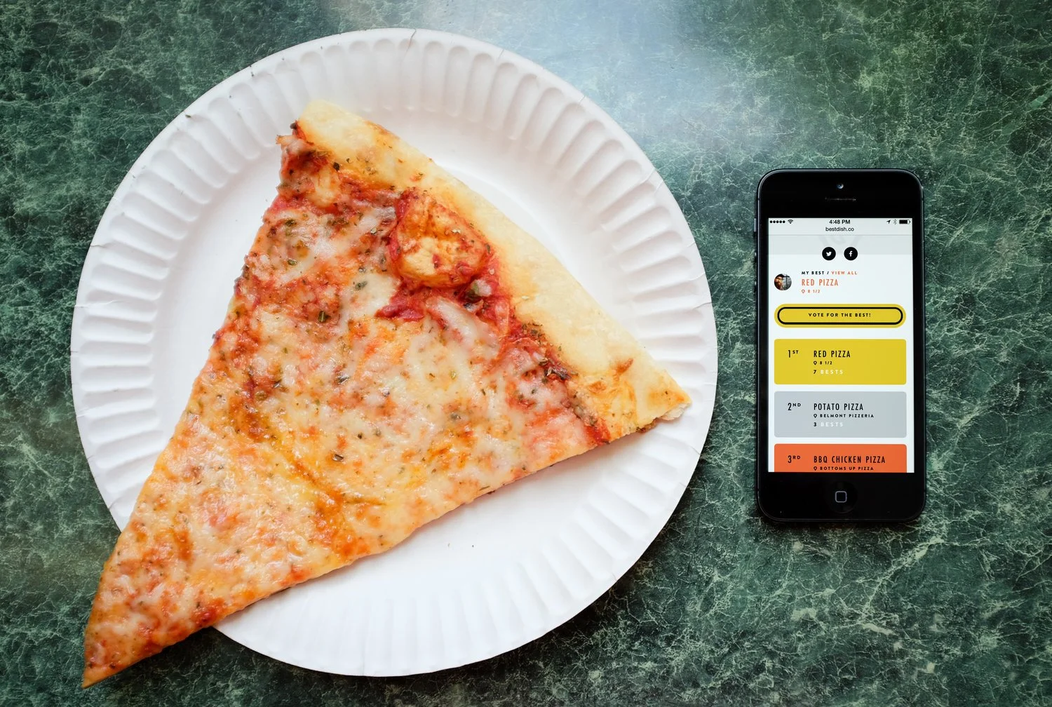A slice of cheese pizza on a white paper plate next to a smartphone displaying a pizza voting app, placed on a green marble surface.