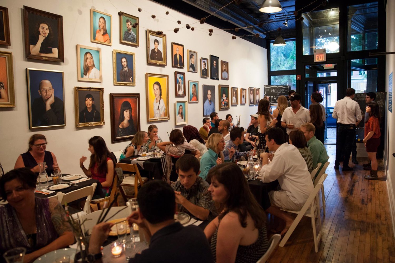 A lively restaurant filled with diners at tables, with a wall decorated with numerous framed portraits of diverse individuals, and large windows at the entrance providing natural light.