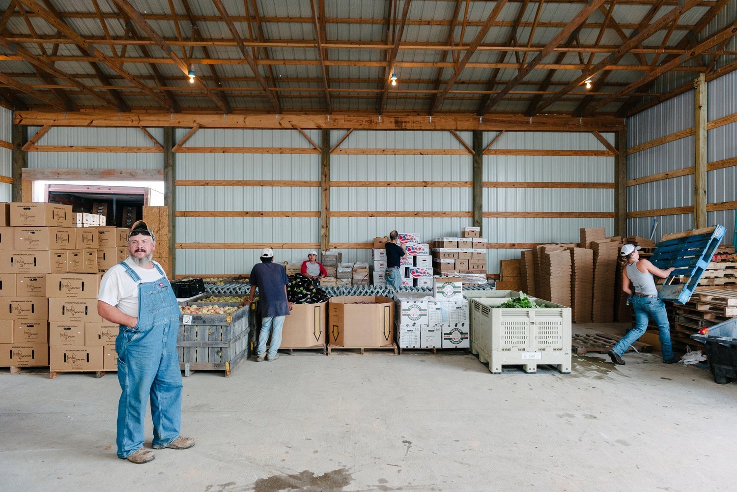 People working inside a warehouse with fresh produce and cardboard boxes, some stacked pallets, and a man standing in the foreground.