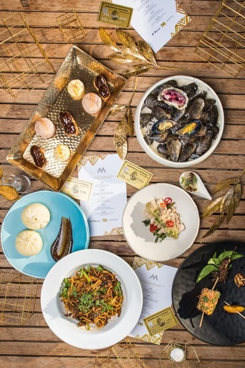 Various Asian dishes including mussels, bao buns, a noodle dish, and appetizers, on a wooden table with gold accents and menus.