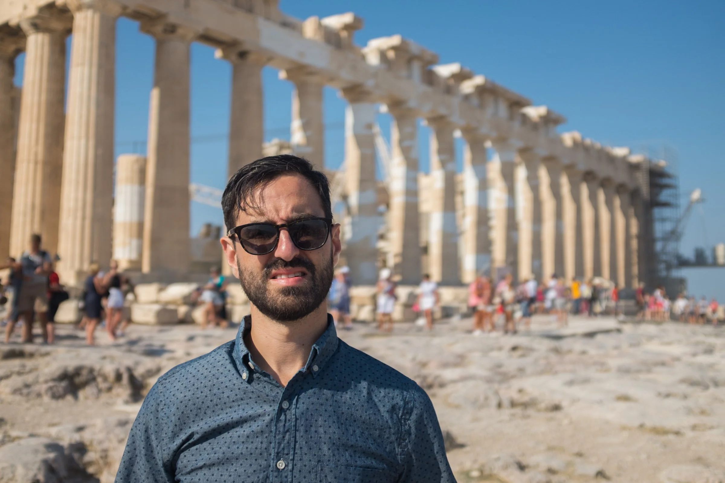 A man with dark hair, a beard, and wearing sunglasses and a blue patterned shirt standing in front of ancient Greek ruins with people walking in the background and a clear blue sky.