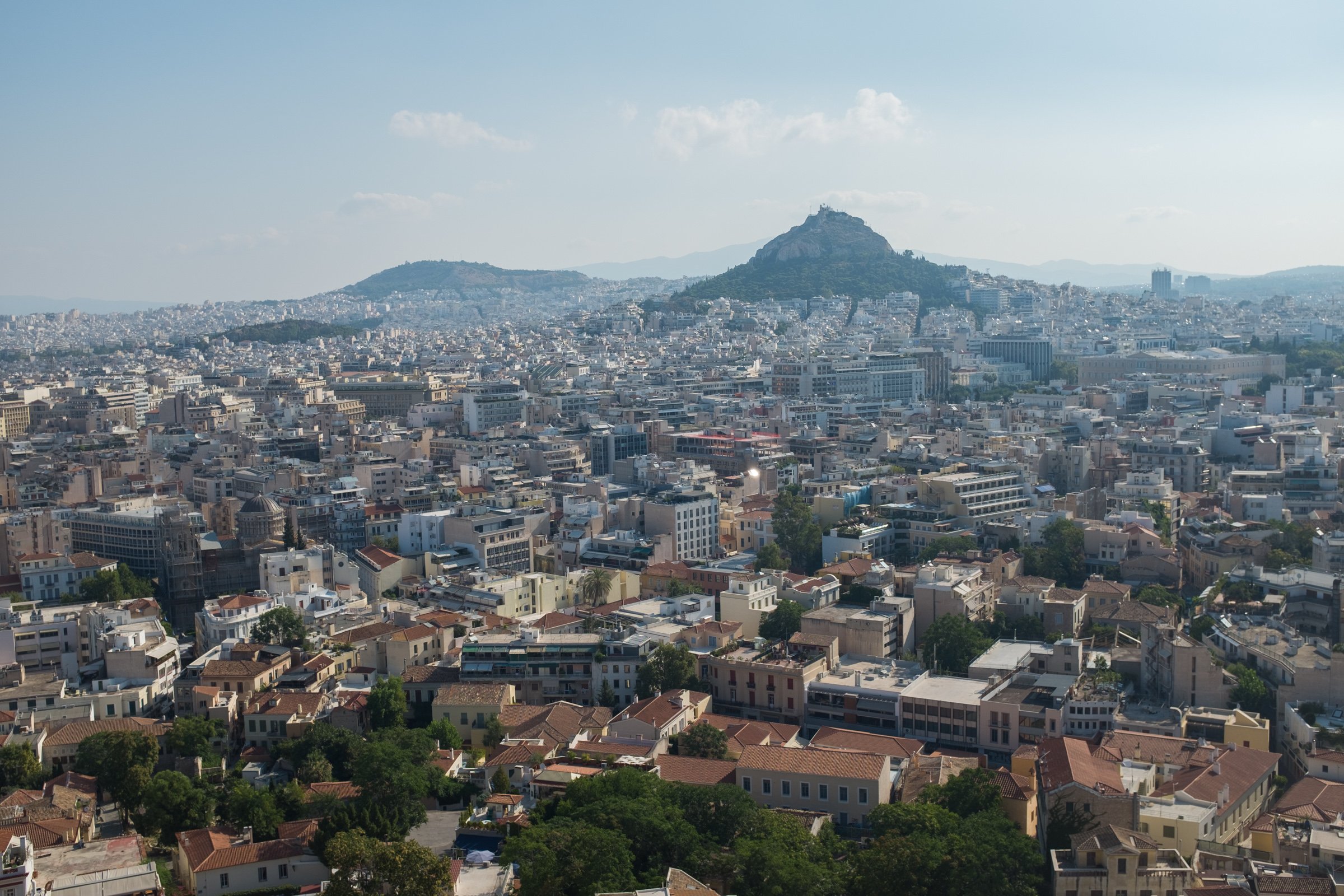 A panoramic view of a cityscape with numerous buildings and houses, with a hill in the background topped by a rocky formation and a few trees, under a clear blue sky with some scattered clouds.