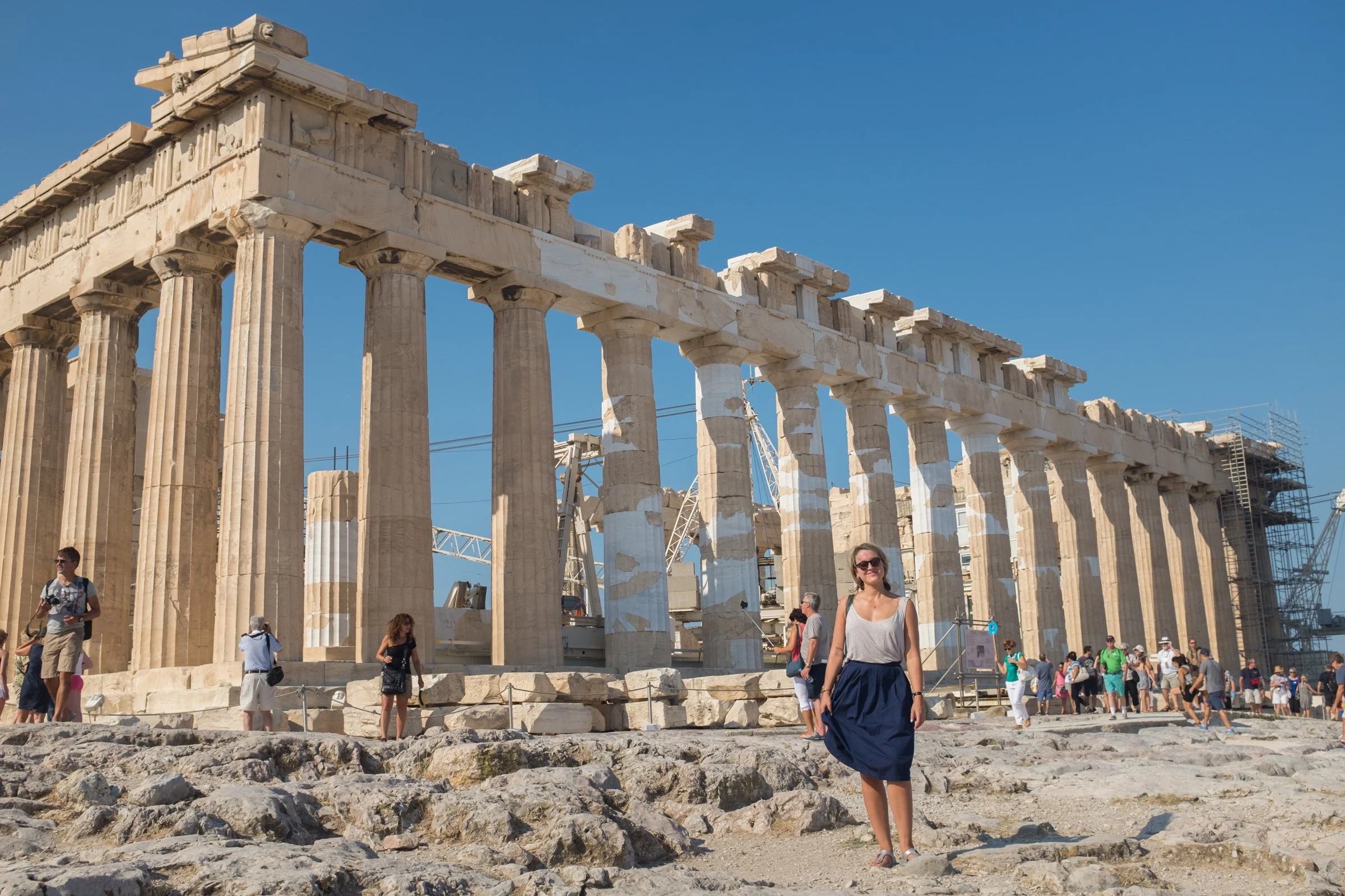 Tourists visiting the Parthenon temple at the Acropolis in Athens, Greece, under a clear blue sky, with some scaffolding and ancient stone ruins in the foreground.