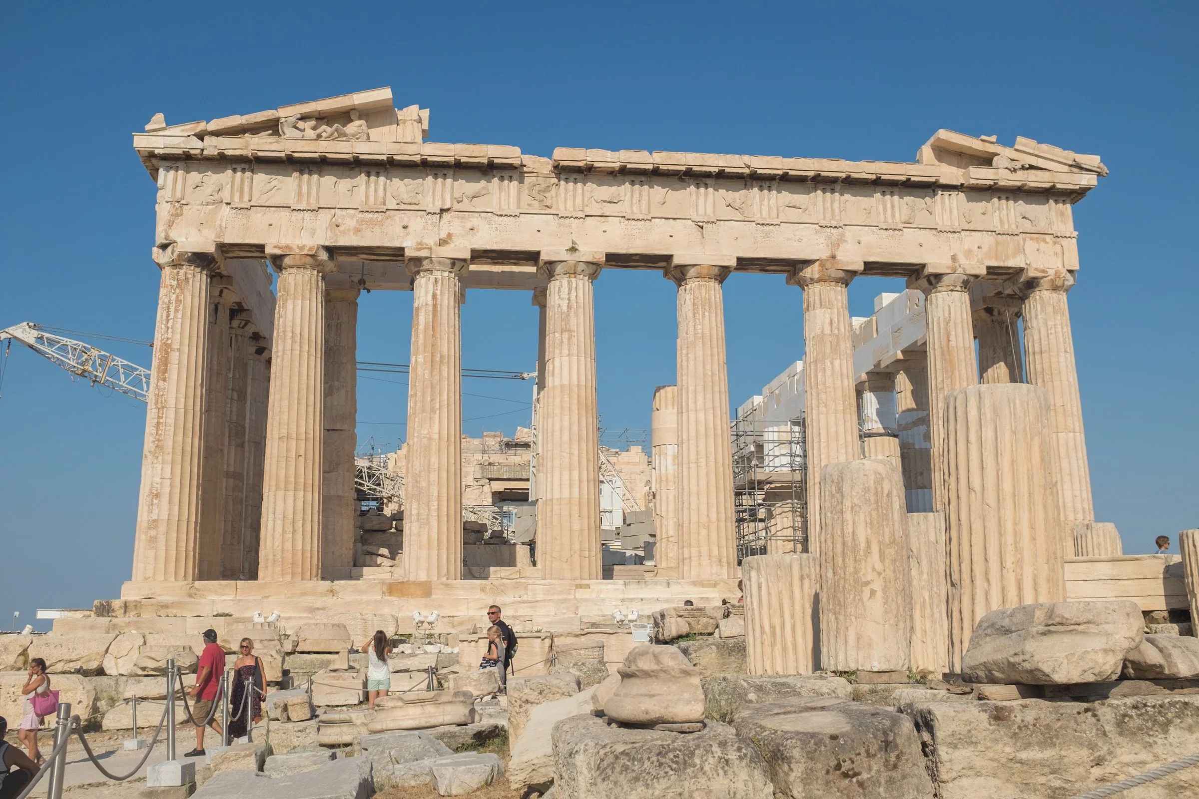 Ancient Greek Parthenon temple ruins with several large columns and a partly damaged entablature against a clear blue sky. Tourists are walking around and exploring the site.