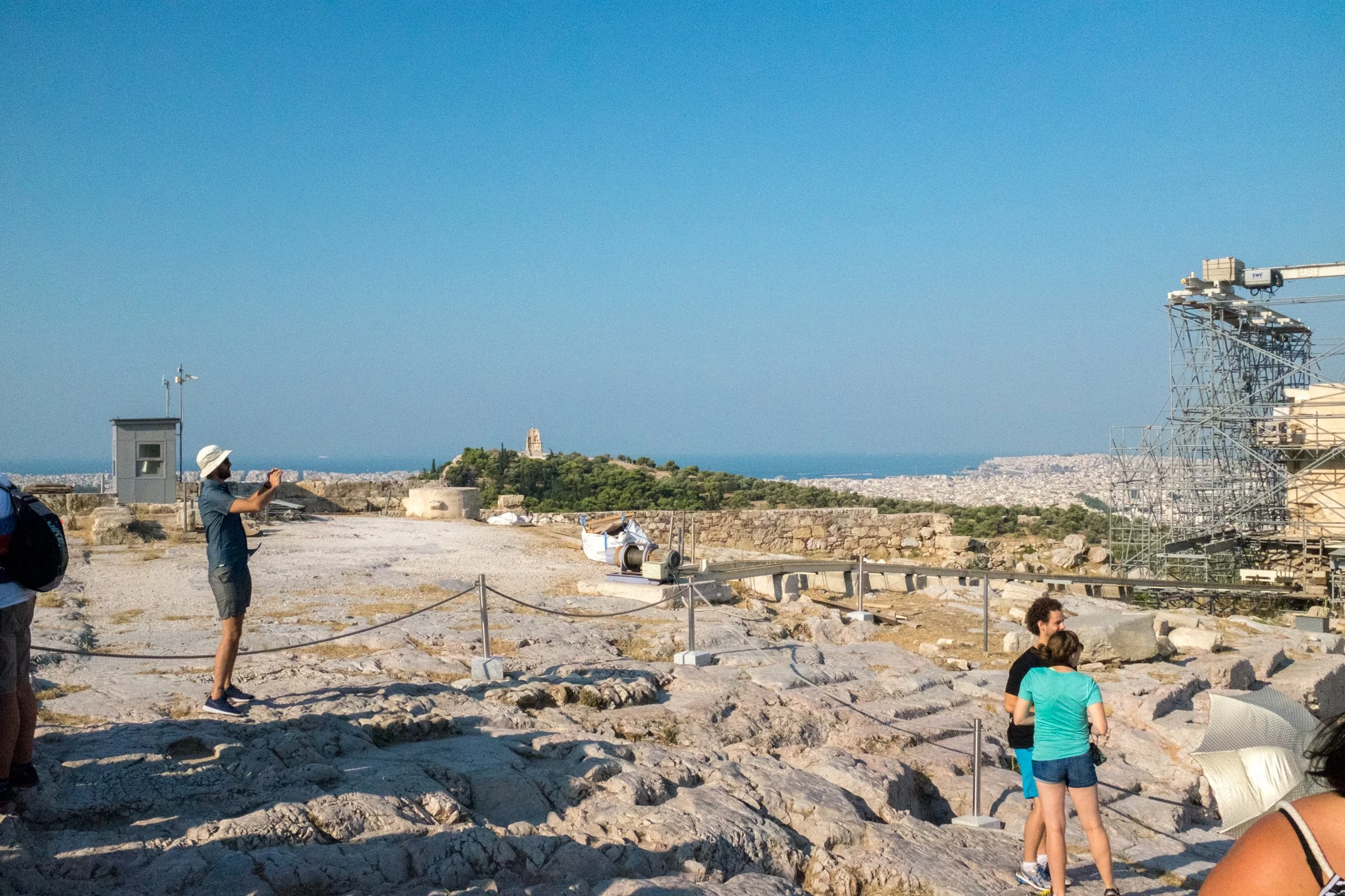 People exploring an archaeological site on a hilltop with scattered ruins, a small building with weather instruments, and a view of the coastline in the background under a clear blue sky.